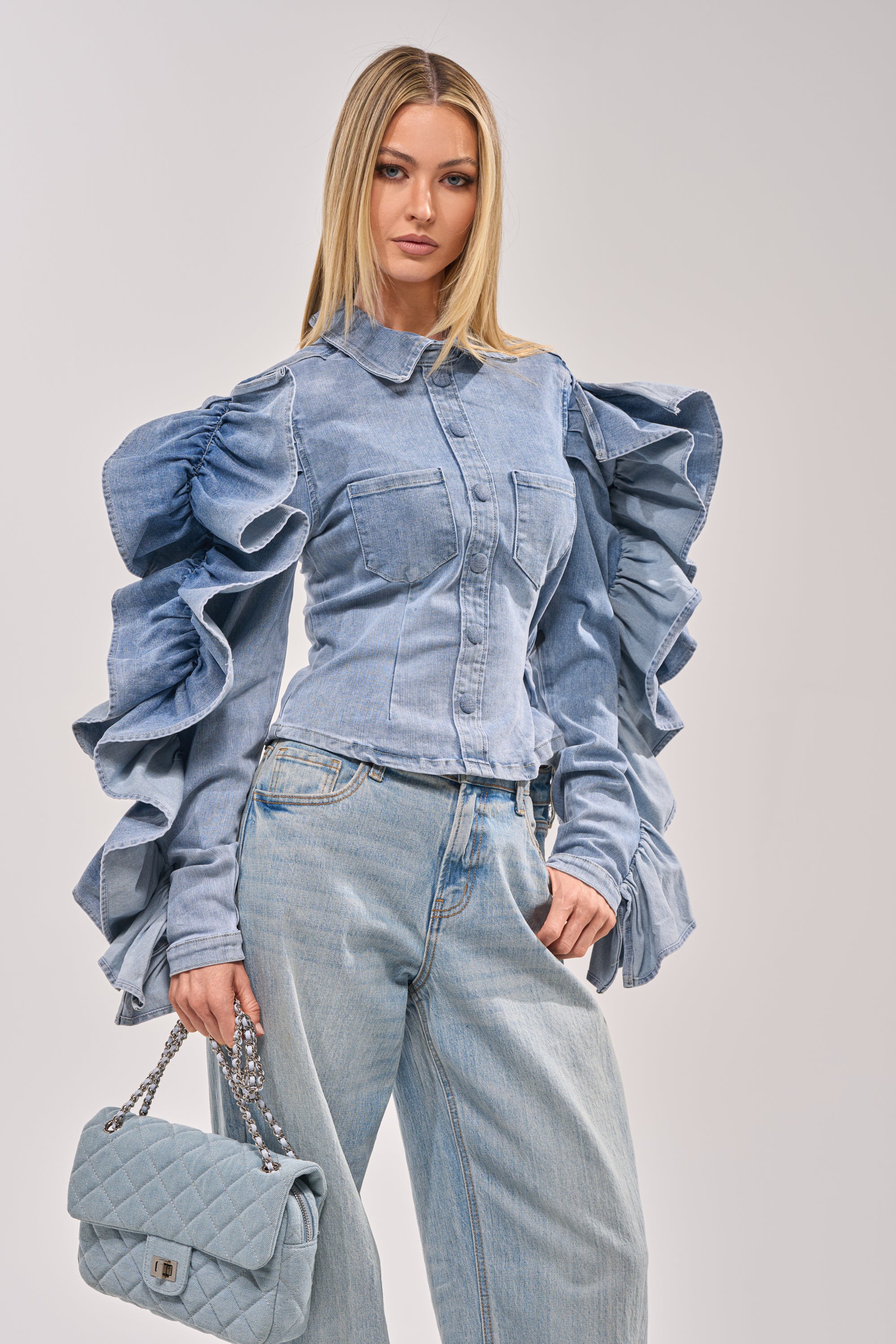 A woman with straight blonde hair and a neutral expression stands against a plain background wearing the DREA RUFFLE SLEEVE DENIM BUTTON DOWN, light wash jeans, and holding a quilted light blue handbag. 2