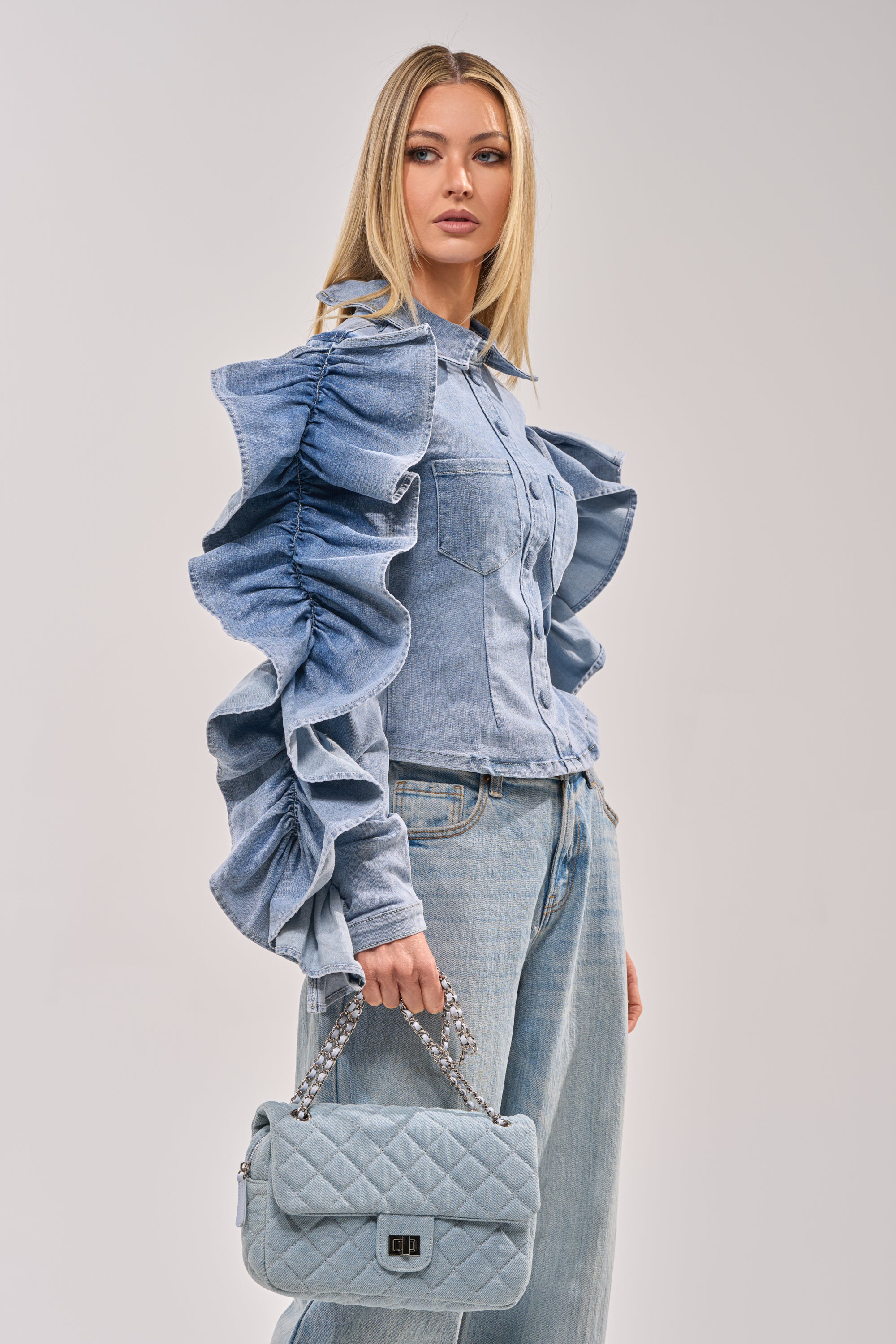 A woman models the DREA RUFFLE SLEEVE DENIM BUTTON DOWN with matching light blue jeans and a quilted chain-strap handbag, turning to the side for a chic brunch look against a plain background. 3