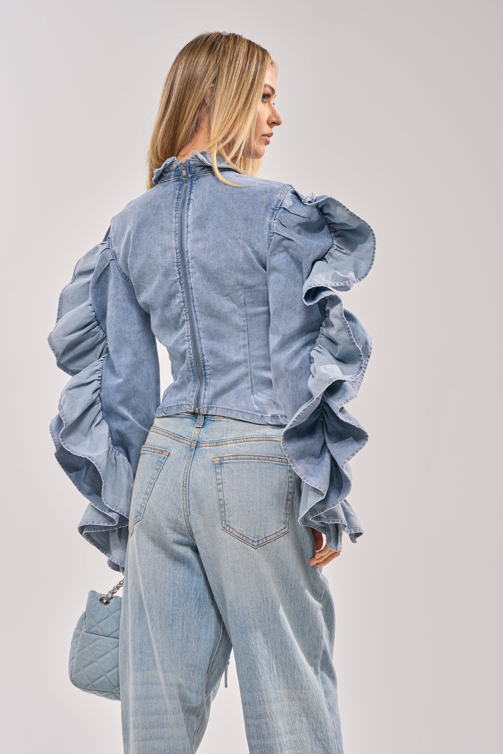 A woman with long blonde hair, facing away, wears the DREA RUFFLE SLEEVE DENIM BUTTON DOWN and light blue jeans. She carries a light blue quilted handbag, ideal for statement brunch looks against a plain background. 4