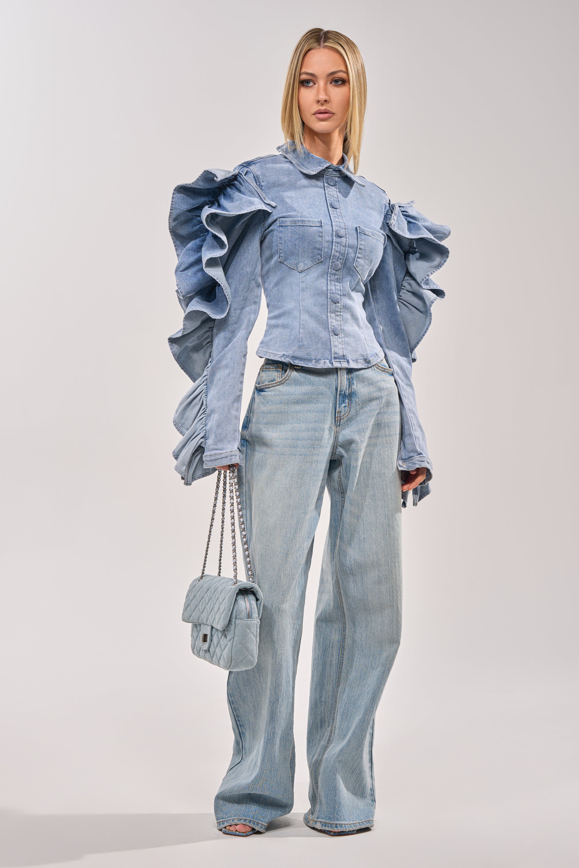 Wearing the DREA RUFFLE SLEEVE DENIM BUTTON DOWN and high-waisted wide-leg jeans, a woman holds a quilted handbag—perfect for statement brunch style—while standing on a plain, light background. 5
