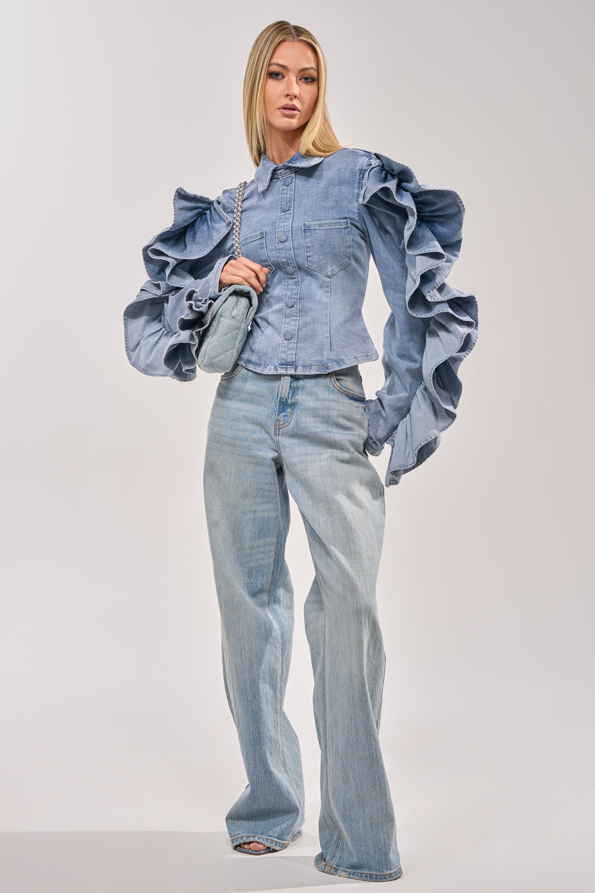 A woman with straight blonde hair stands against a plain background wearing the DREA RUFFLE SLEEVE DENIM BUTTON DOWN and light blue wide-leg jeans, carrying a small shoulder bag for standout brunch style. 6
