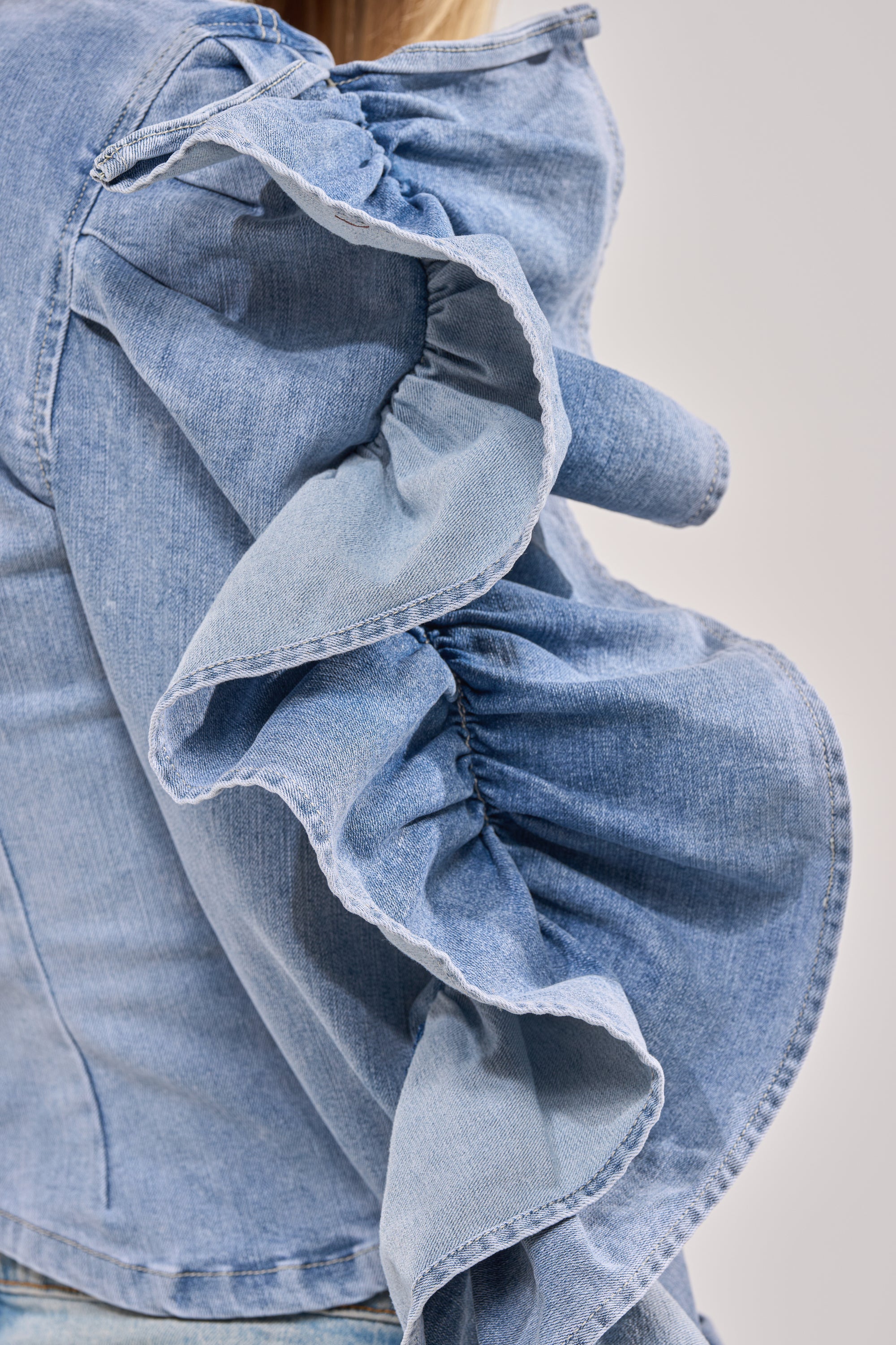 Close-up of a person wearing the DREA RUFFLE SLEEVE DENIM BUTTON DOWN, featuring detailed stitching and textured fabric—perfect for making a statement at brunch against a neutral background. 7