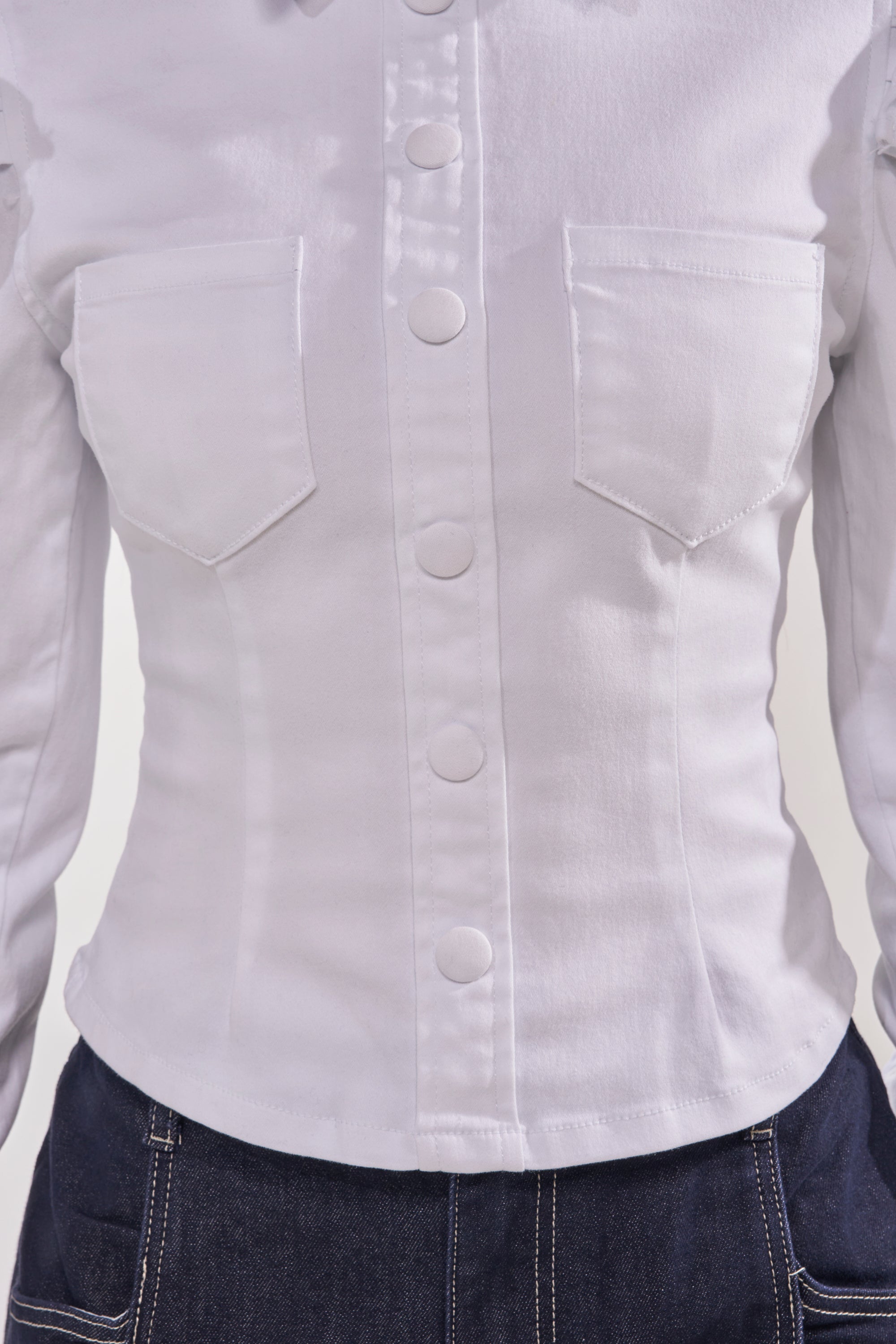 A close-up of a person wearing the DREA RUFFLE SLEEVE BUTTON DOWN under a white denim jacket, paired with dark blue jeans with white stitching for a chic, polished office look. 10