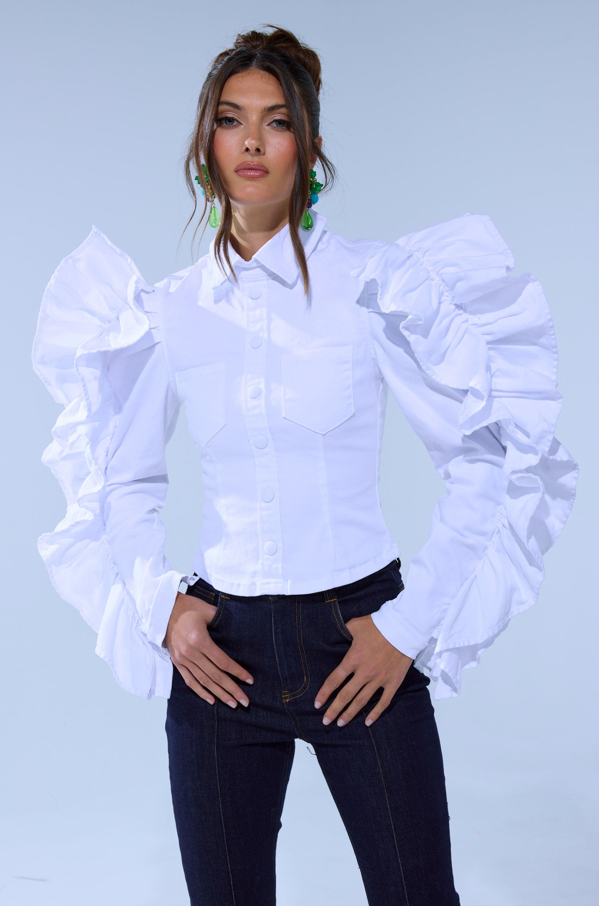 A woman poses in the DREA RUFFLE SLEEVE BUTTON DOWN, a tailored cotton-blend shirt with large ruffled puff sleeves, paired with dark jeans and green statement earrings, her hair styled up against a plain light background.