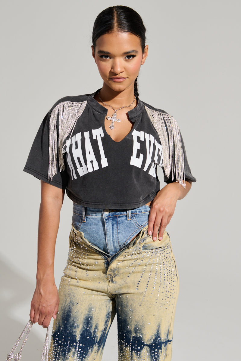 A person stands confidently, sporting the KISS IT SHORT SLEEVE RHINESTONE FRINGE TEE with "WHAT EVER" emblazoned on it. They flaunt high-waisted, two-tone jeans with a distressed look and a hint of cotton-blend comfort. With a small bag in hand and a necklace adorned with decorative elements, their style is effortlessly chic.
