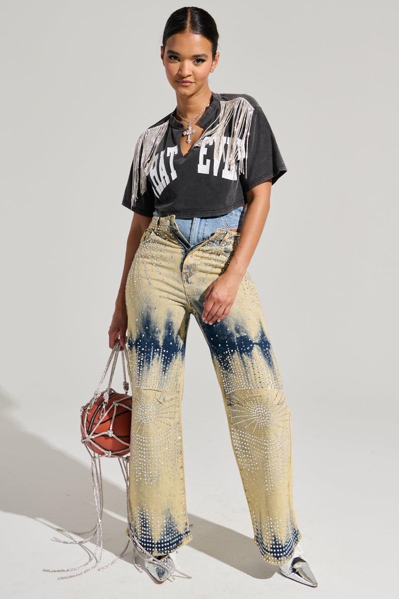 A person poses confidently in the KISS IT SHORT SLEEVE RHINESTONE FRINGE TEE, a cotton-blend top with bold "WHAT EVER" lettering. They wear high-waisted tie-dye jeans featuring white patterns and pair them with metallic silver shoes. Completing the look is a beaded bag shaped like a basketball.