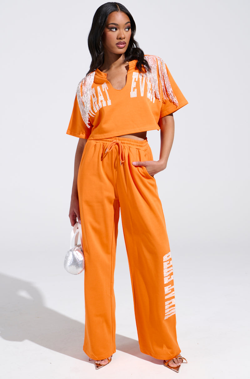 A woman stands confidently clad in high-rise "Whatever You Want Rhinestone" sweatpants, displaying text down one leg. Her outfit, with fringed orange shoulders and a boldly printed top, contrasts elegantly with her silver bag and heels against the plain white backdrop.