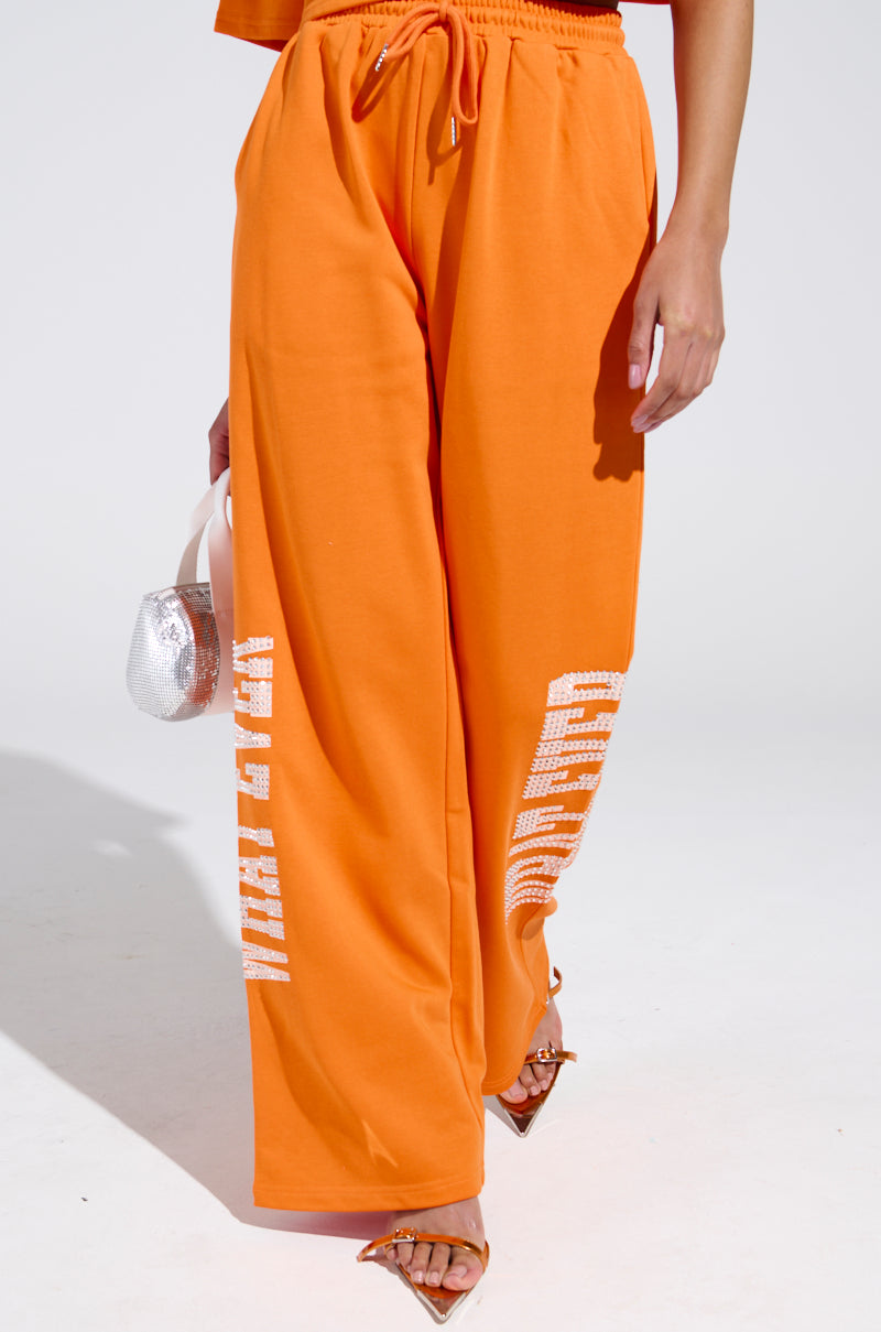 A person in bright orange "WHATEVER YOU WANT RHINESTONE SWEATPANTS" with white text on one leg holds a small silver handbag, paired with an elasticized waistband top and pointed-toe heels against a plain white background.