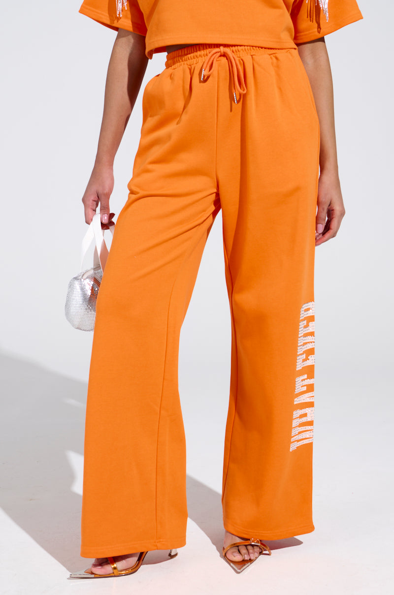 A person wearing high-rise WHATEVER YOU WANT RHINESTONE SWEATPANTS in bright orange with white side lettering stands against a plain background, holding a small metallic bag and donning orange-heeled sandals. The focus is on the outfit from waist to feet.