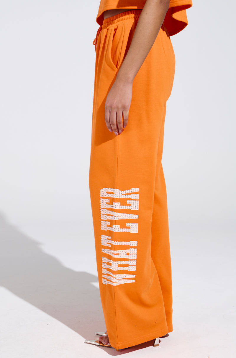 Wearing vibrant orange WHATEVER YOU WANT RHINESTONE SWEATPANTS with "Character" in white down the side, paired with an orange cropped top, this ensemble pops against a plain white background.