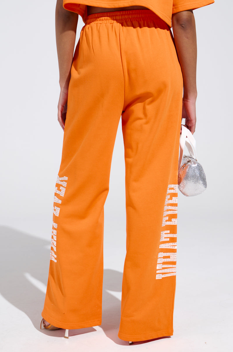 A person in bright orange WHATEVER YOU WANT RHINESTONE SWEATPANTS, featuring white lettering and an elastic waistband, pairs the look with a small silver handbag. The outfit stands out against the plain, light-colored background.