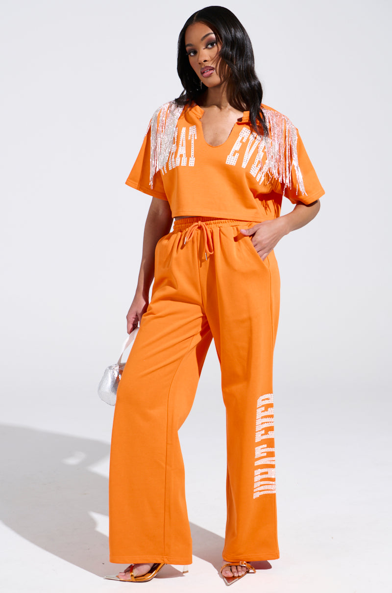 A person confidently wears WHATEVER YOU WANT RHINESTONE SWEATPANTS, featuring an elastic waistband for comfort. The ensemble includes a top with white lettering and silver fringe on the shoulders, paired with a small silver bag. Their heels strikingly contrast the white background.