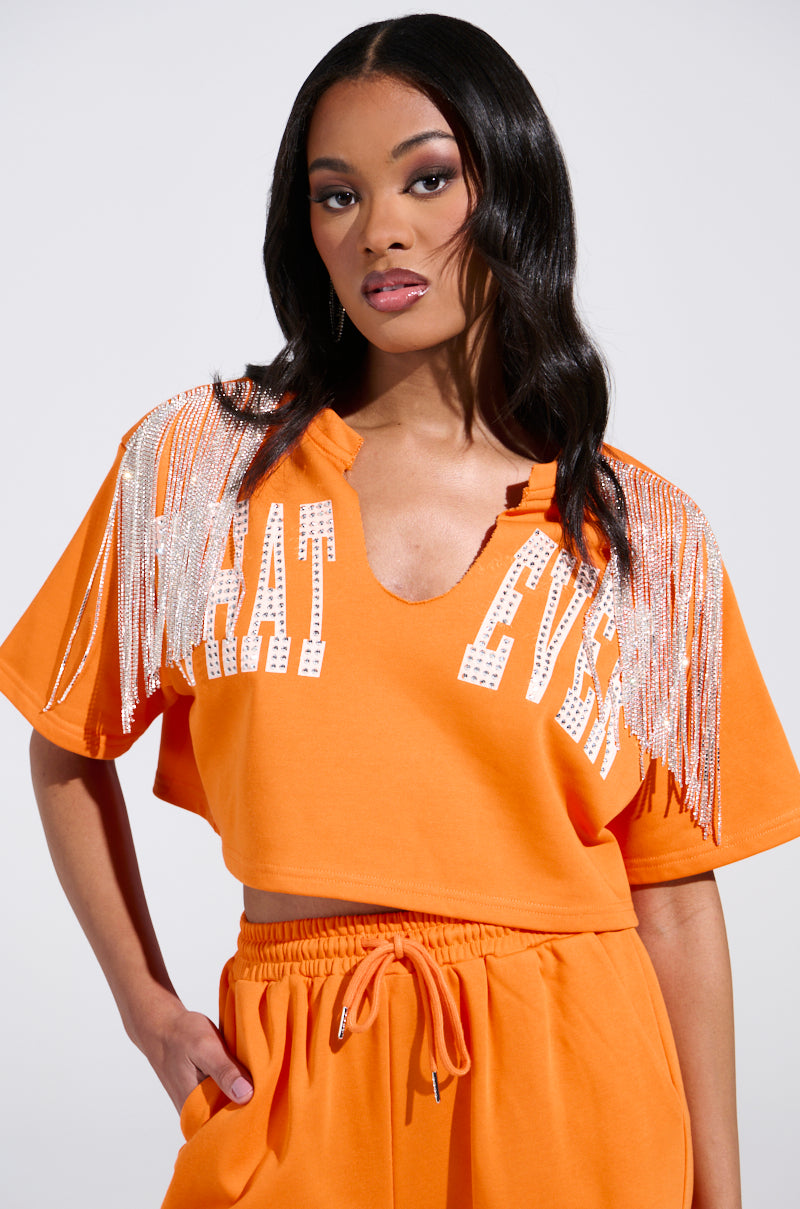 A person with long dark hair poses against a plain background, exuding effortless style in an orange WHATEVER YOU WANT CROPPED RHINESTONE FRINGE TEE, adorned with rhinestone fringes on the shoulders and "WHAT EVER" printed on the stretchy sweatshirt top.