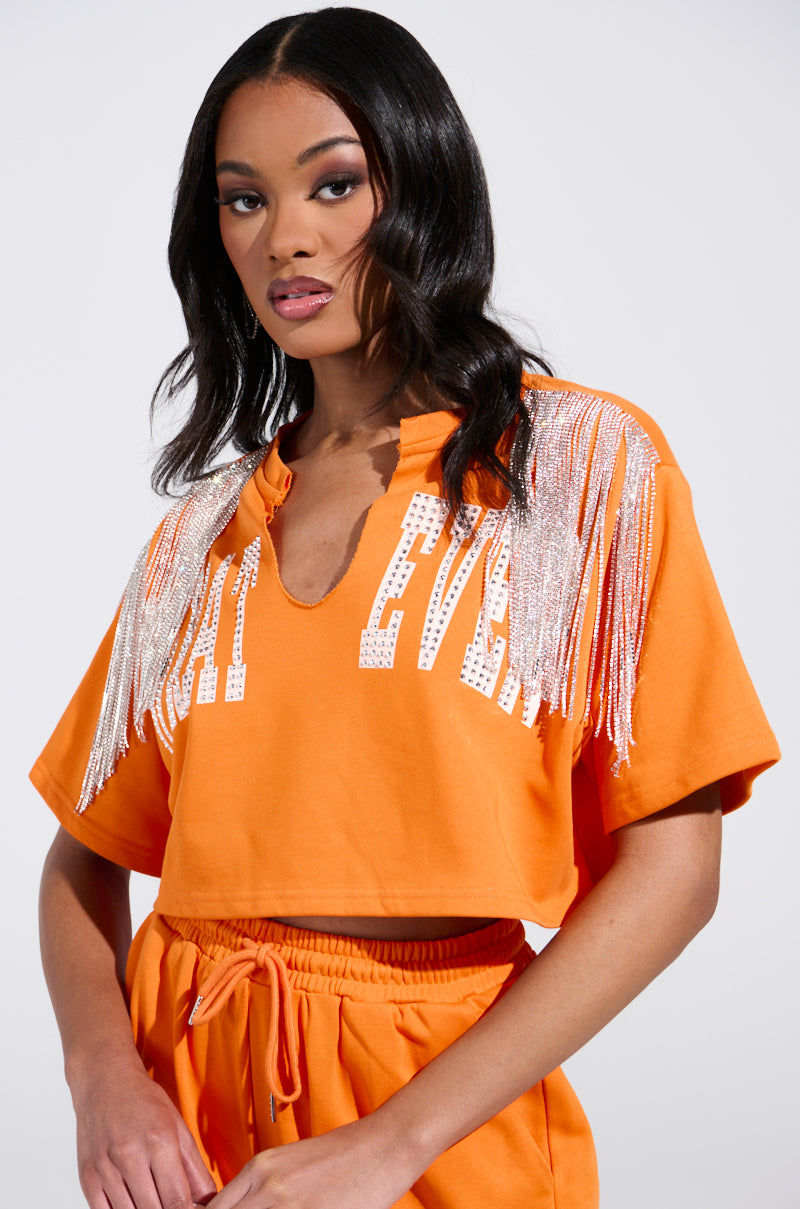 The person dons an orange ensemble, highlighted by the WHATEVER YOU WANT cropped rhinestone fringe tee with sparkling embellishments and sequined text. The V-neck top pairs seamlessly with drawstring pants as their wavy hair cascades while they gaze confidently at the camera.