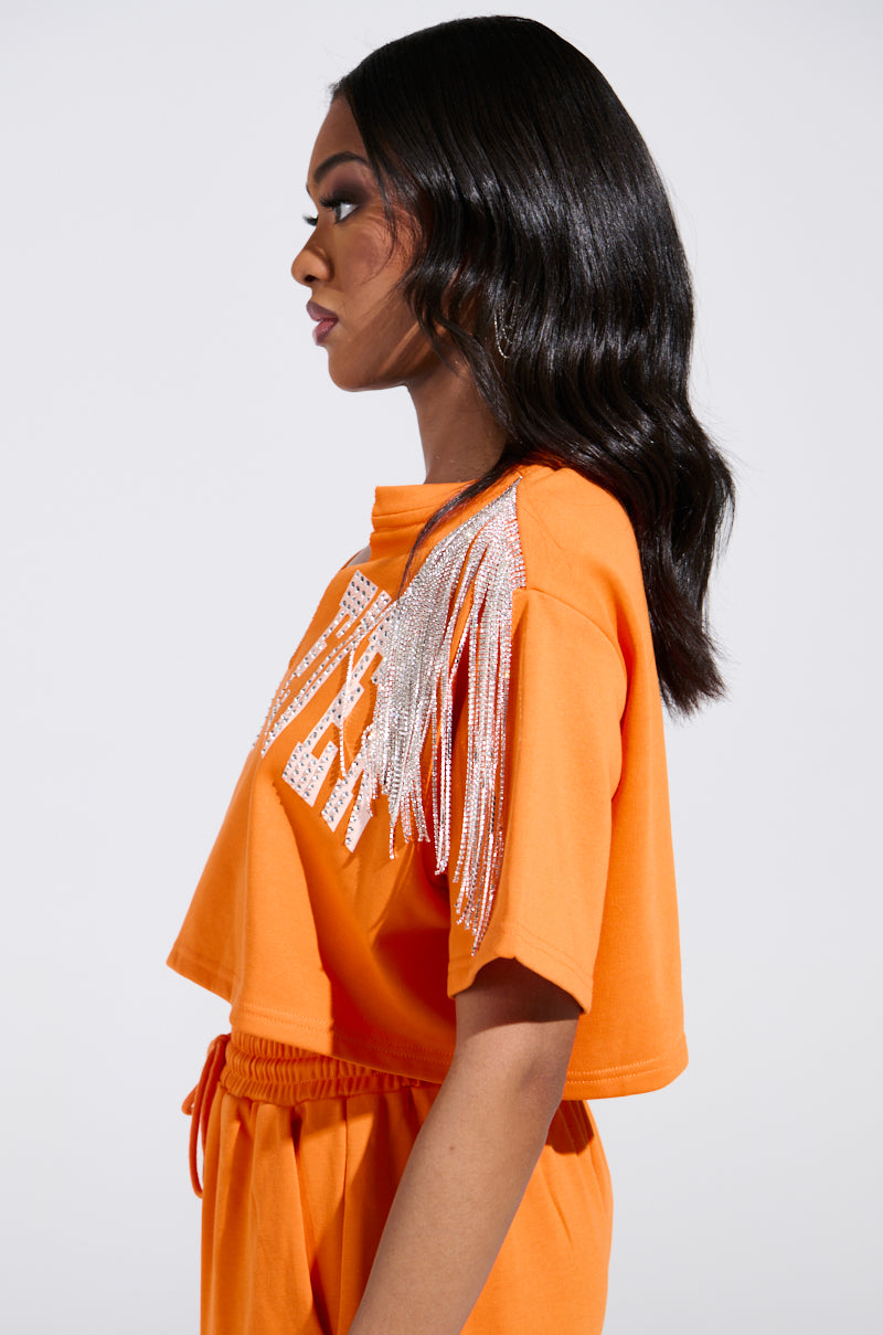 A person with long black hair poses sideways in an orange WHATEVER YOU WANT CROPPED RHINESTONE FRINGE TEE against a plain, light-colored background.