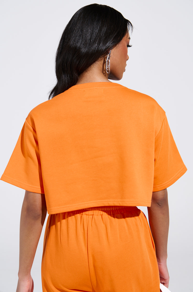 A person with long dark hair is wearing the WHATEVER YOU WANT CROPPED RHINESTONE FRINGE TEE in orange and matching high-waisted pants. They are facing away from the camera against a plain, light background.