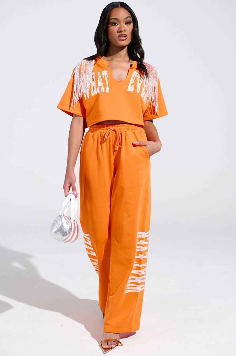 A woman models a chic orange two-piece set featuring the WHATEVER YOU WANT CROPPED RHINESTONE FRINGE TEE paired with a stylish small silver bag and strappy sandals. Her confident pose pops against the plain white background.