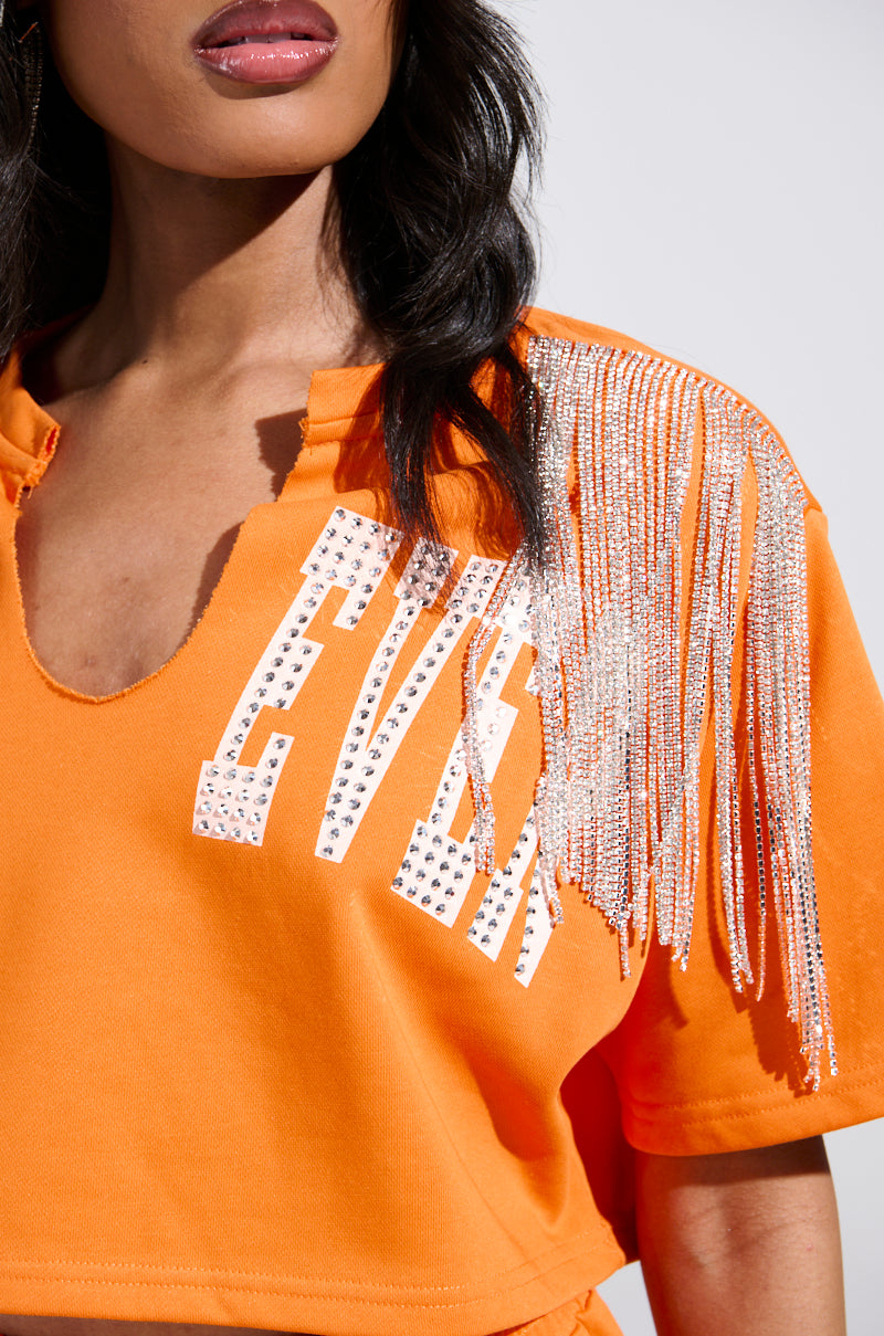 A person with long dark hair and a neutral expression wears the WHATEVER YOU WANT CROPPED RHINESTONE FRINGE TEE in orange, featuring "EVER" in dazzling rhinestones and sparkling fringe embellishments on the shoulders.