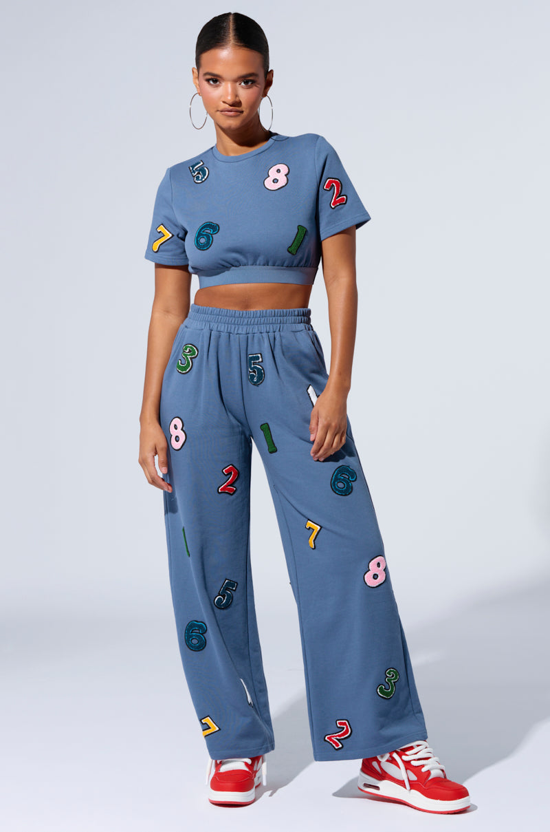 A person stands confidently in the COUNT IT UP! WIDE LEG JOGGER set, featuring a coordinated blue crop top and wide-leg pants adorned with colorful numbers. They complete their look with red and white sneakers and large hoop earrings, posing against a plain light background.