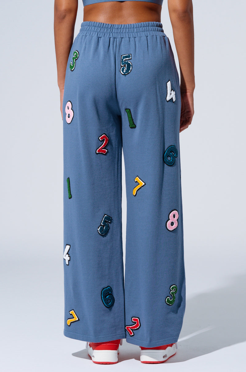 A person stands with their back to the camera, showcasing the COUNT IT UP! WIDE LEG JOGGER in blue. The jogger is decorated with an array of colorful, embroidered numbers scattered across the fabric in various colors and sizes, adding a playful and vibrant charm. Complementing the joggers, the person sports red and white sneakers.