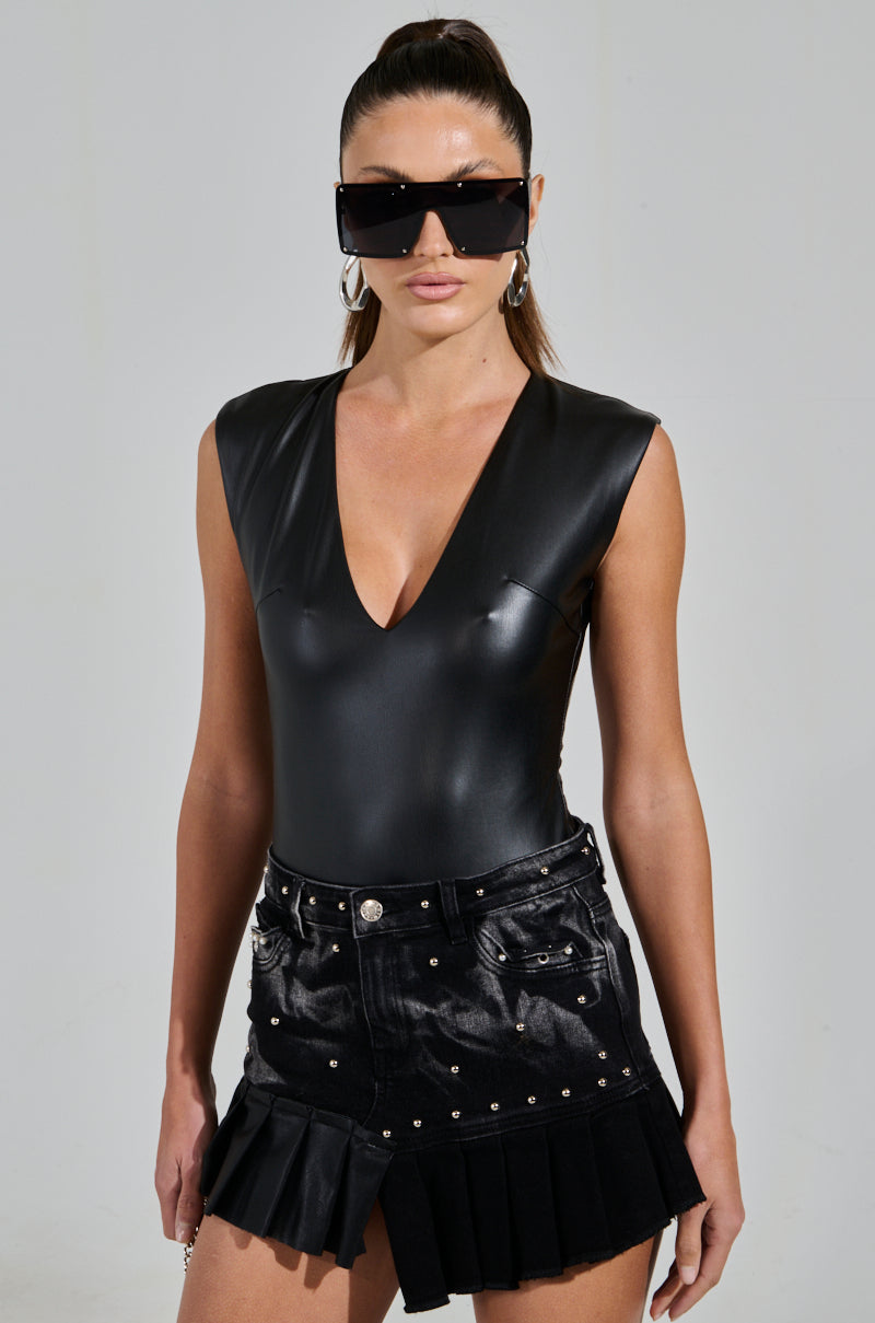 A person with long hair styled high wears large sunglasses and a JOELLE FAUX LEATHER SLEEVELESS PLUNGE BODYSUIT in sleek black, paired with a black skirt adorned with metallic studs and pleated details, highlighting their curve-hugging silhouette.