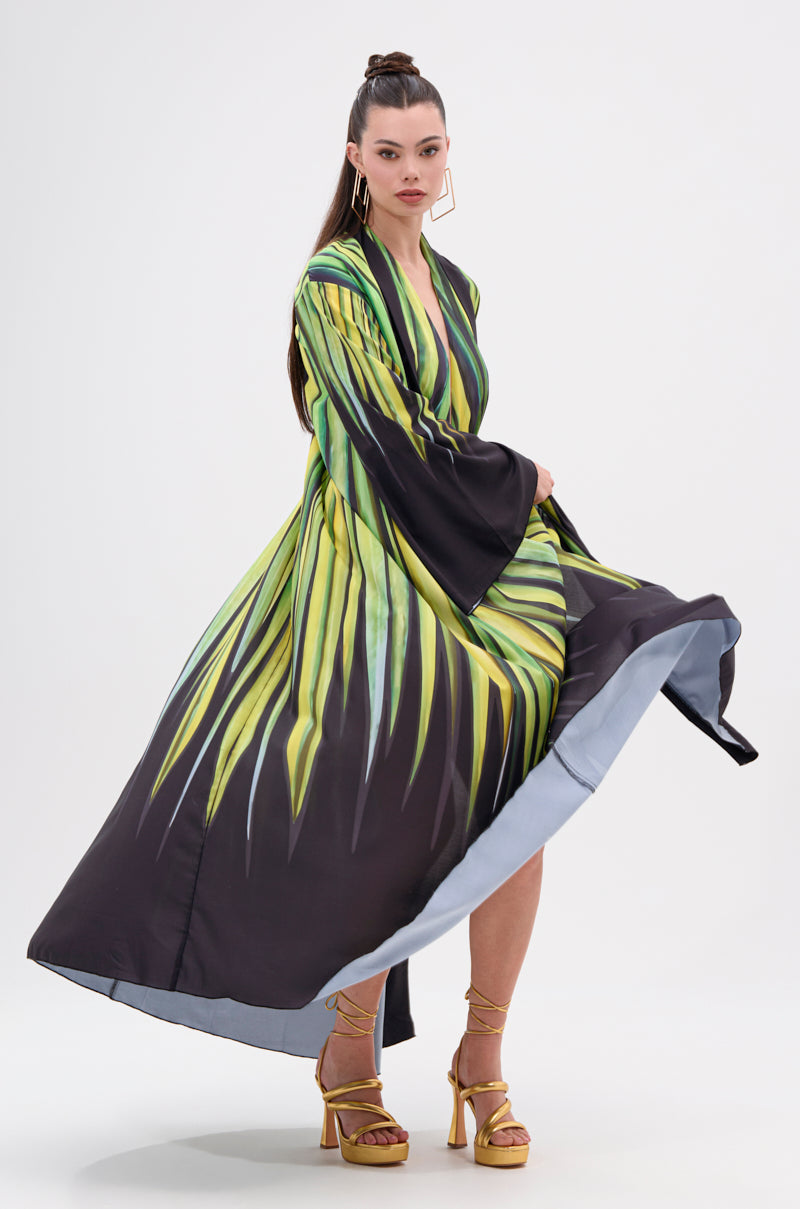Wearing the DEEP IN THE WOODS DUSTER with a green and yellow abstract graphic on satin, she pairs it with gold heels. Her long hair is tied back, adding movement as the flowing robe turns with her.