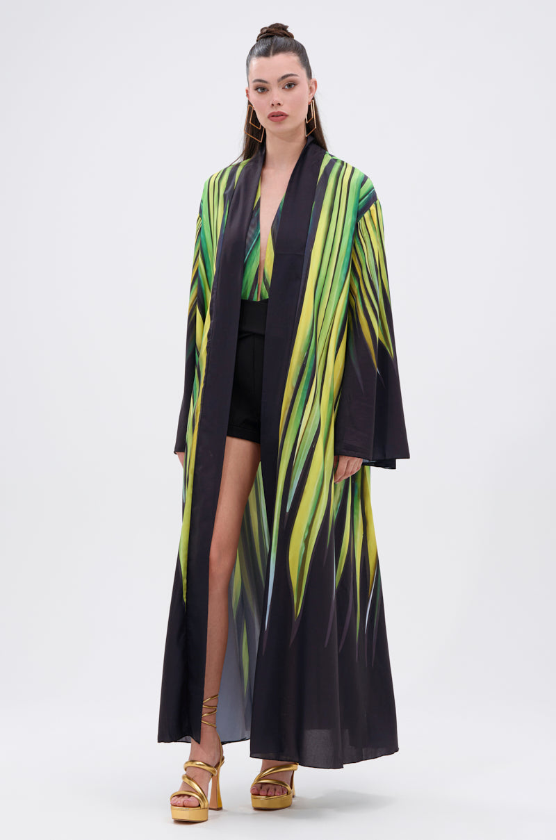 A woman with long hair in a high ponytail faces forward, wearing the DEEP IN THE WOODS DUSTER with green and yellow leaf-like patterns over a green top, black shorts, and gold sandals.
