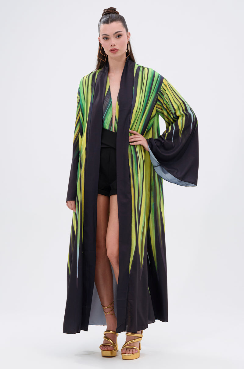 A woman in the DEEP IN THE WOODS DUSTER, a long black satin kimono with green and yellow abstract print, styled with a matching top, high-waisted black shorts, and gold strappy heels stands against a plain white background.
