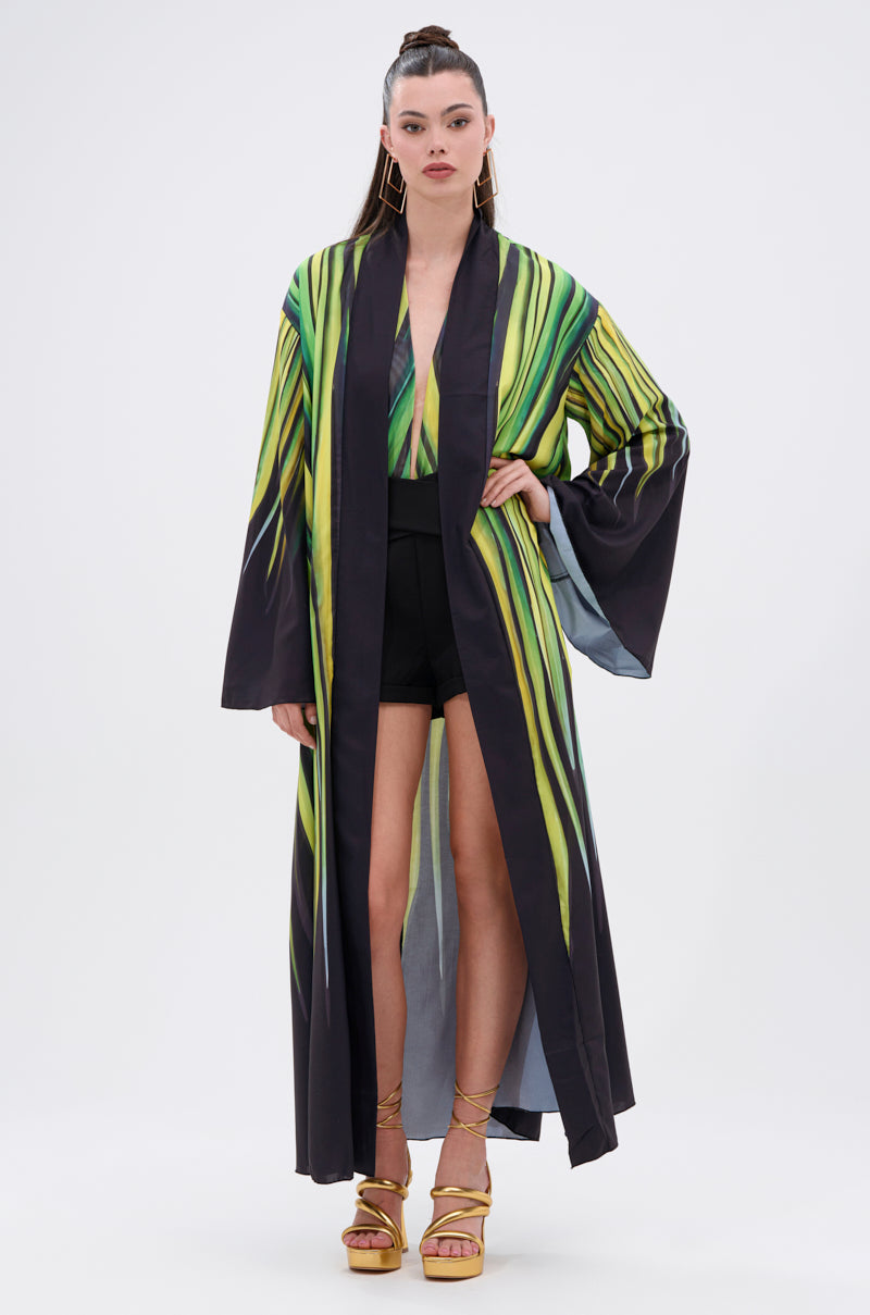A woman stands facing forward in the DEEP IN THE WOODS DUSTER featuring a green and yellow abstract print, layered over a matching top and black shorts. She pairs it with yellow high-heeled sandals and wears her hair in a high ponytail.