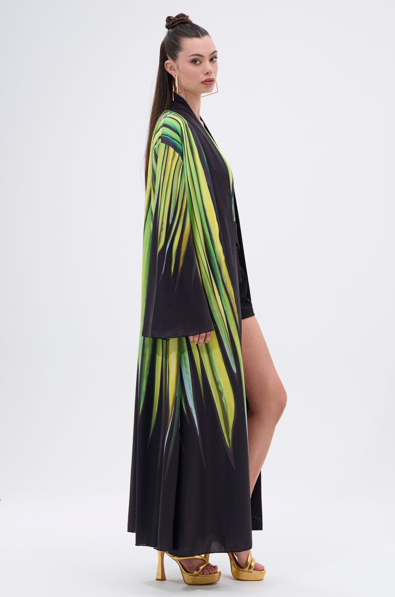 A woman with long brown hair stands sideways against a white background, wearing the DEEP IN THE WOODS DUSTER—a satin robe with an abstract print—over a black outfit and gold high heels.