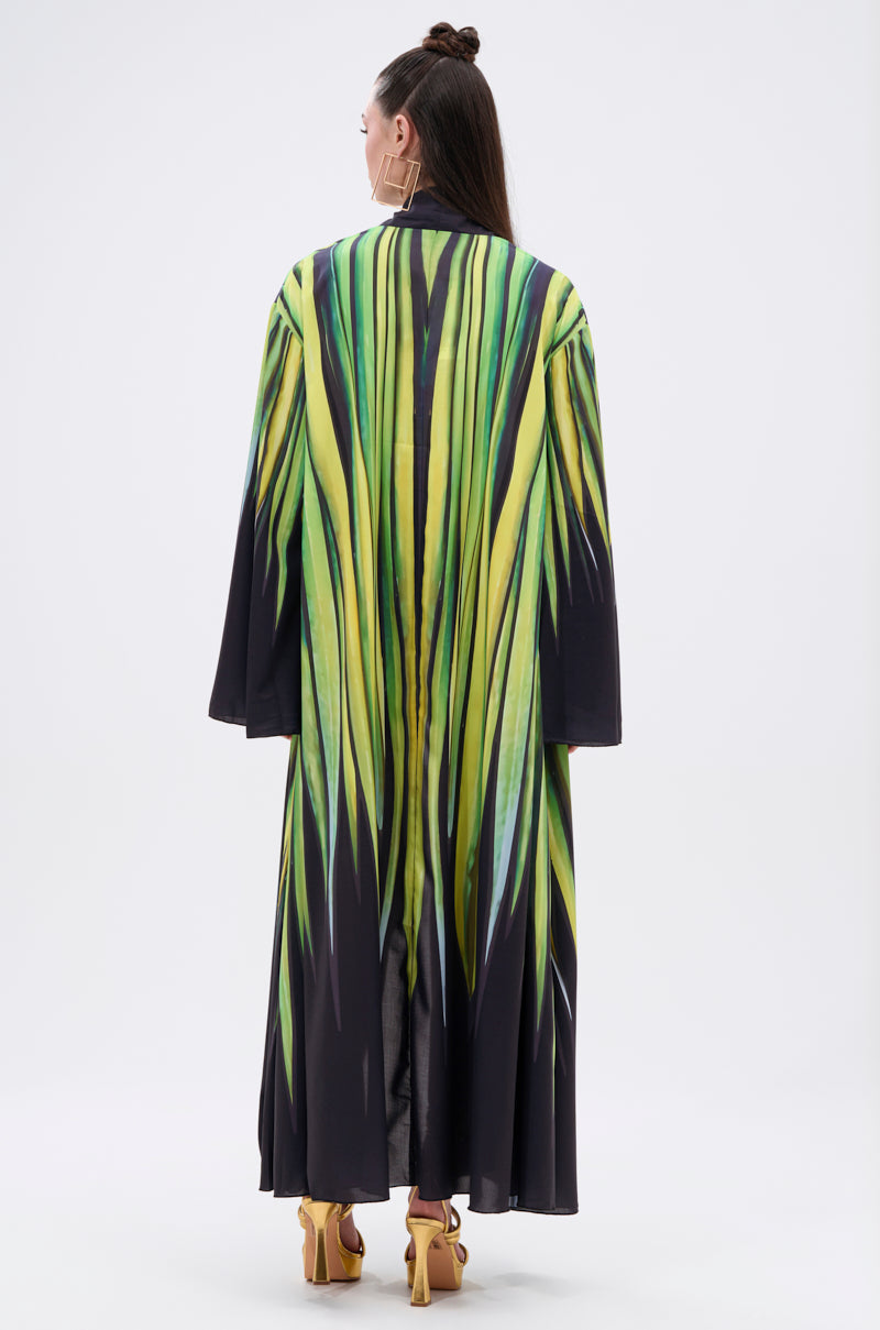 A person stands with their back to the camera, wearing the DEEP IN THE WOODS DUSTER—a long satin coat featuring a bold green and yellow abstract print—with their brown hair tied up and paired with yellow high-heeled sandals.