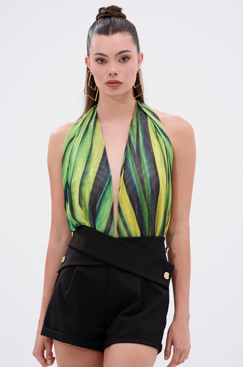 A woman wears the DEEP IN THE WOODS BODYSUIT, featuring a plunging halter neckline and green-yellow abstract print, paired with high-waisted black shorts with gold buttons, a high bun, and large geometric earrings. 0