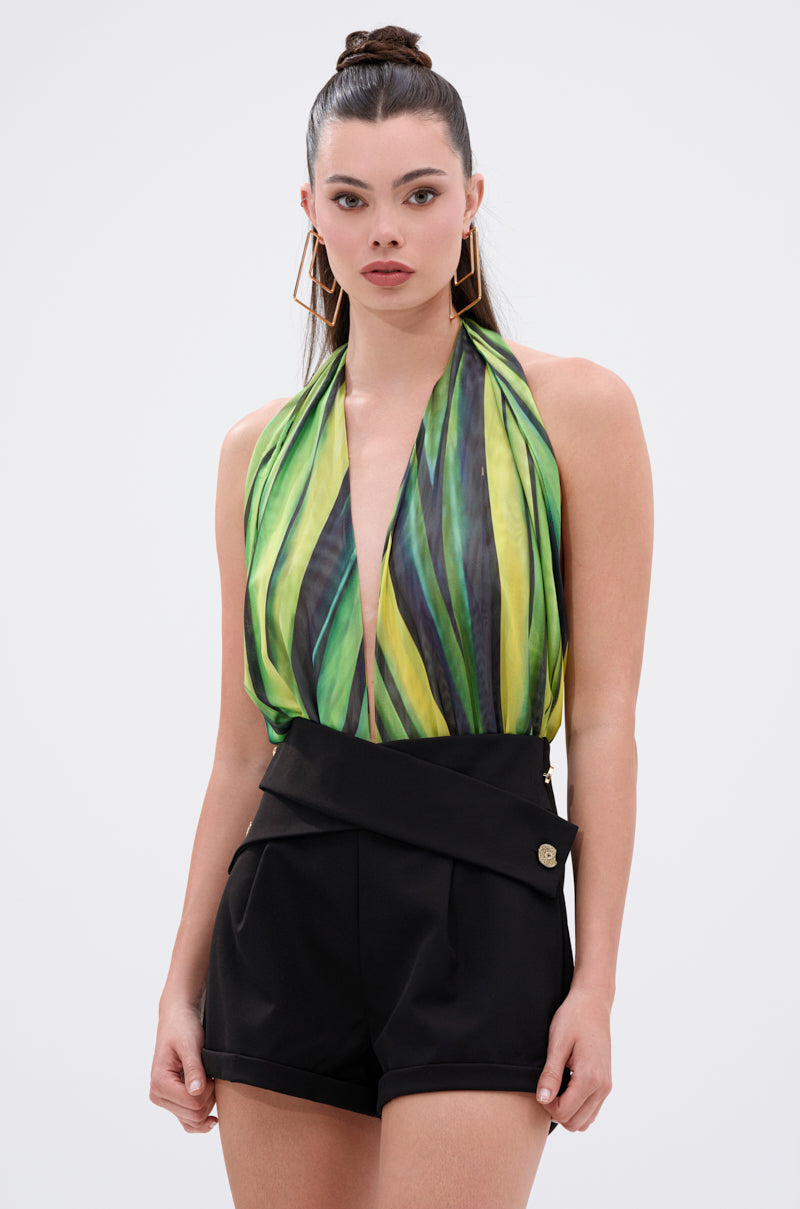 A woman with straight hair in a high bun and large geometric gold earrings poses against a plain background, wearing the DEEP IN THE WOODS BODYSUIT with a plunging halter neckline and green/yellow abstract print, paired with high-waisted black shorts. 1