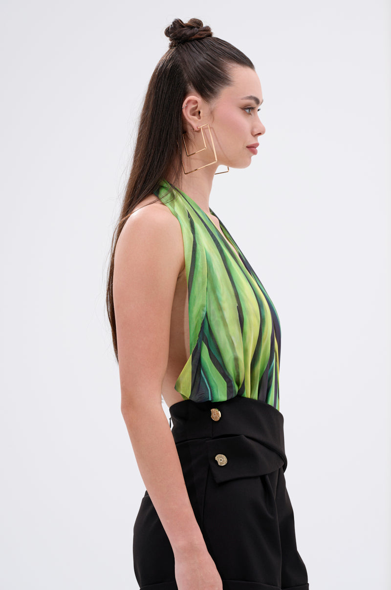A woman with long brown hair styled half-up wears large square earrings, high-waisted black pants with gold buttons, and the DEEP IN THE WOODS BODYSUIT—a green piece with an abstract print and plunging halter neckline—standing in profile. 2