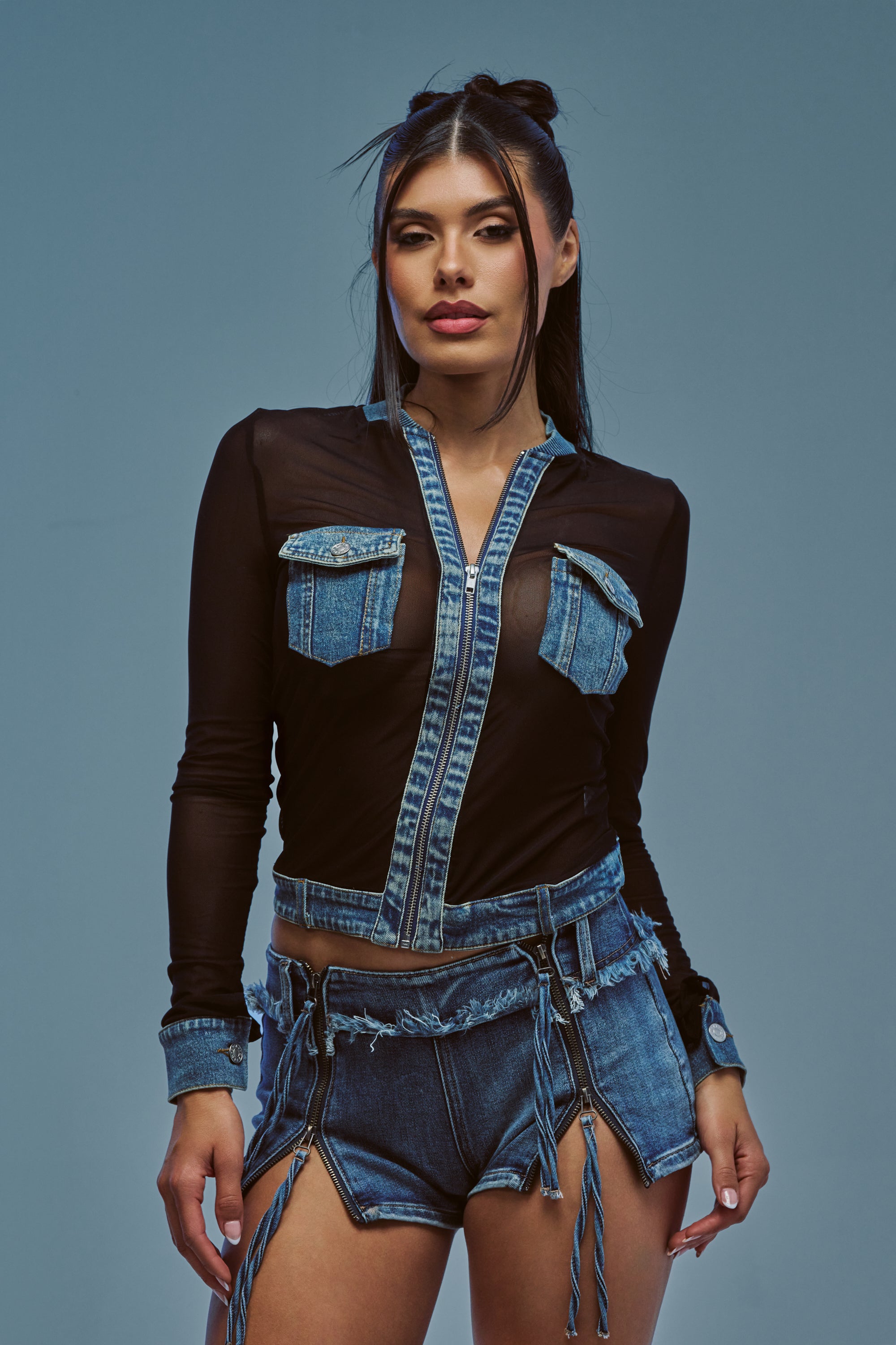 Confidently posing against a plain blue background, the woman showcases effortless street style in the DENIM TRIM MESH ZIP UP TOP paired with matching denim shorts.
