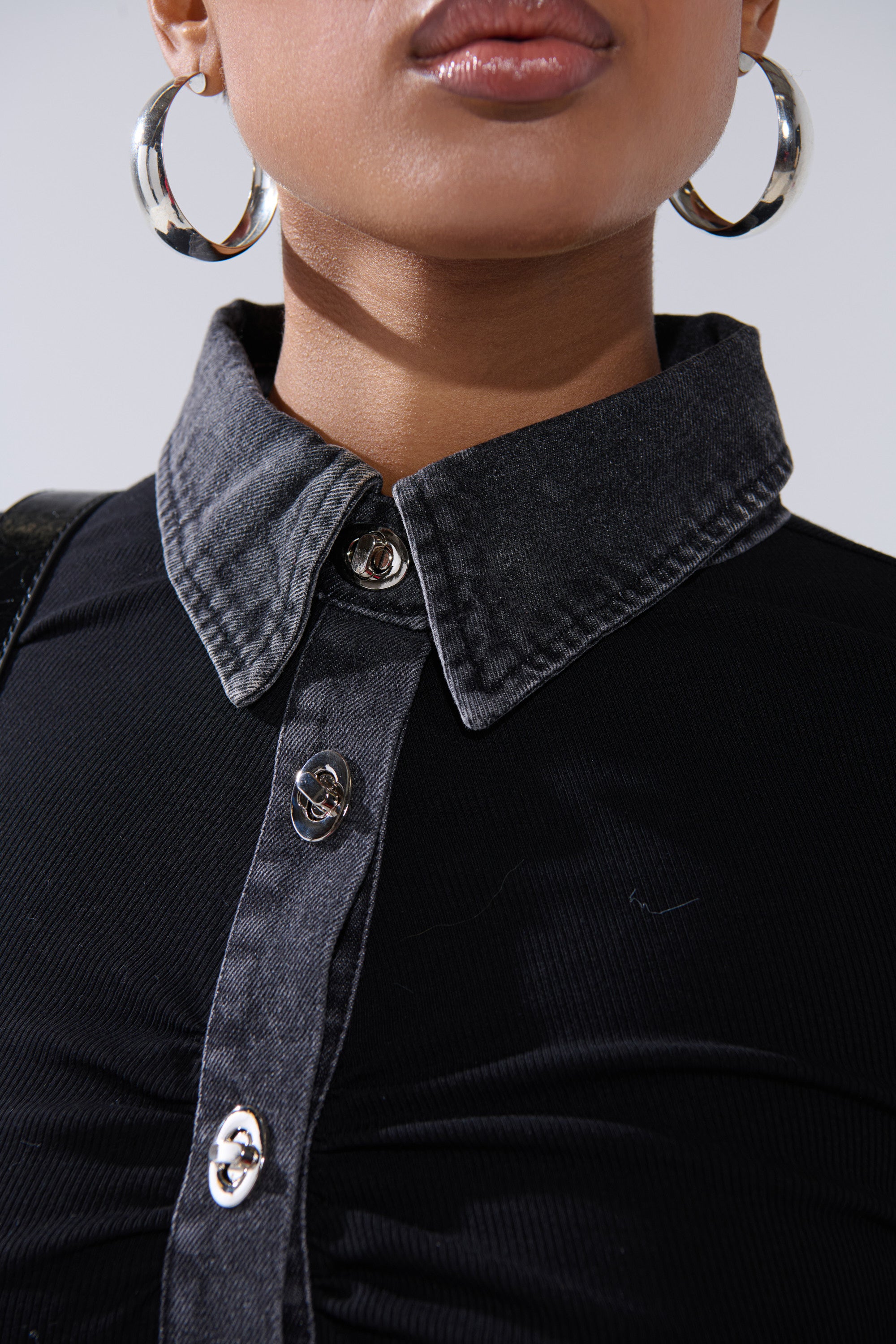 A close-up of a person wearing the SHANNON KNIT BUTTON DOWN, highlighting its prominent buttons and subtle denim-trimmed collar. The lower face and neck are partially visible; paired with large silver hoop earrings.