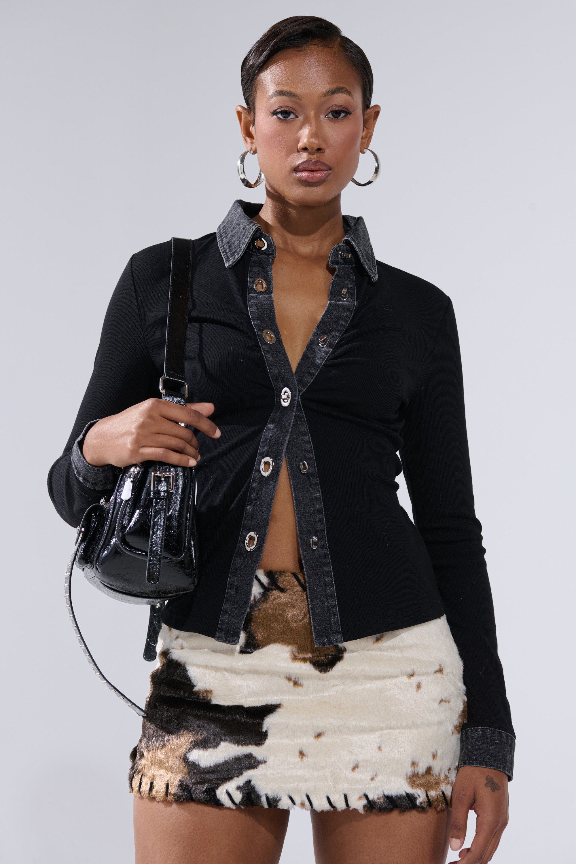 A woman with short hair wears the SHANNON KNIT BUTTON DOWN featuring a denim trim collar, paired with a black and white fuzzy mini skirt, large hoop earrings, and a small black handbag. She stands against a plain light background.