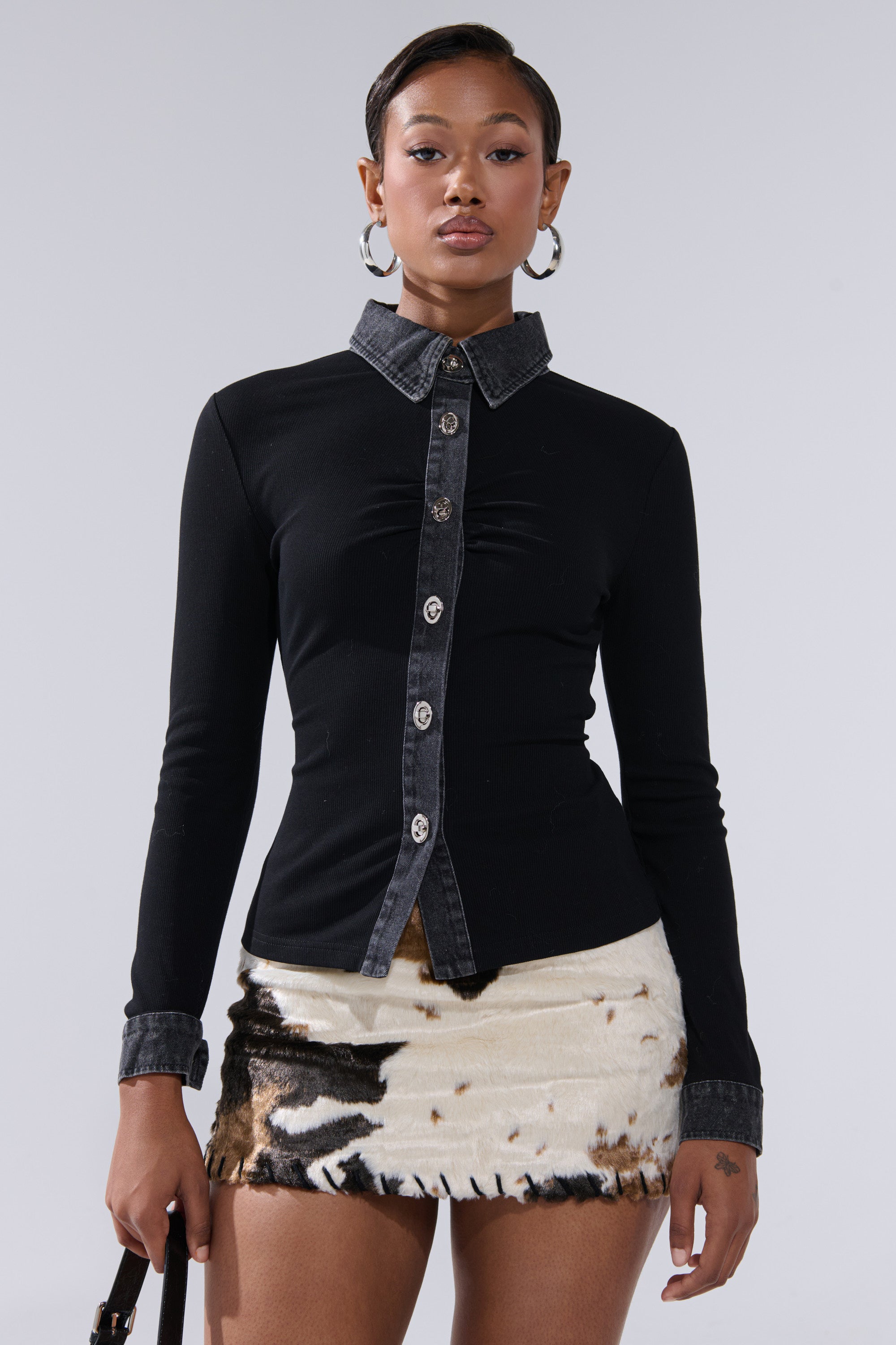 A woman stands confidently in the SHANNON KNIT BUTTON DOWN, featuring denim trim on the collar and cuffs, styled with a short animal-print skirt. She accessorizes with hoop earrings and a black handbag against a plain background.