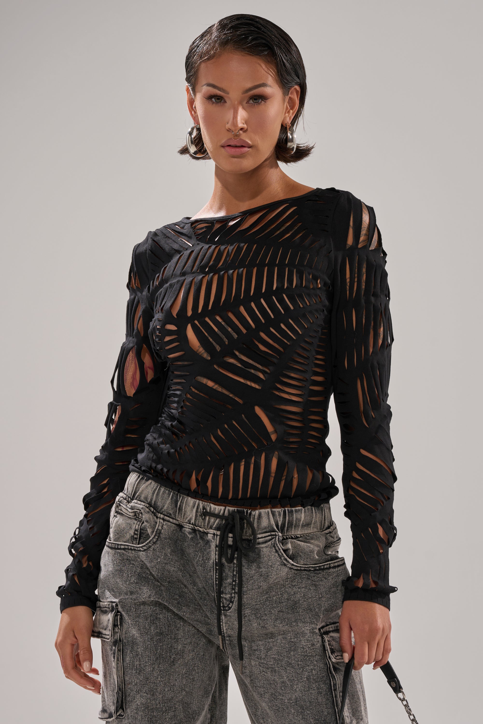 A woman stands facing forward against a plain light gray background, wearing the SPIDER WEB CUTOUT TOP—black, long-sleeve with spider web cutouts and a nude underlayer—paired with gray baggy drawstring waist jeans.