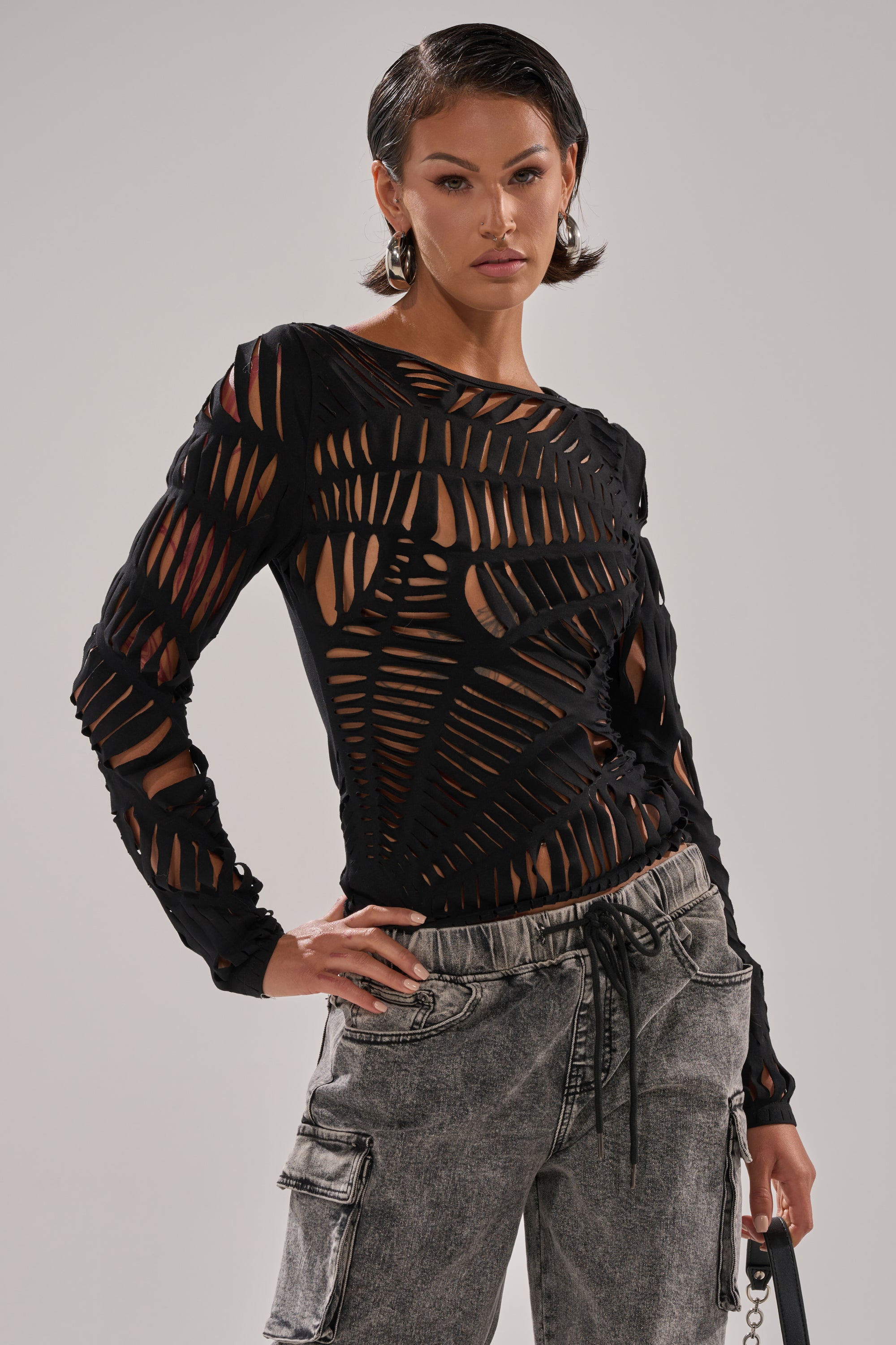 A woman with short dark hair models the SPIDER WEB CUTOUT TOP, which features geometric patterns and shredded cut details. She pairs it with light gray cargo pants and poses with one hand on her hip against a plain background.