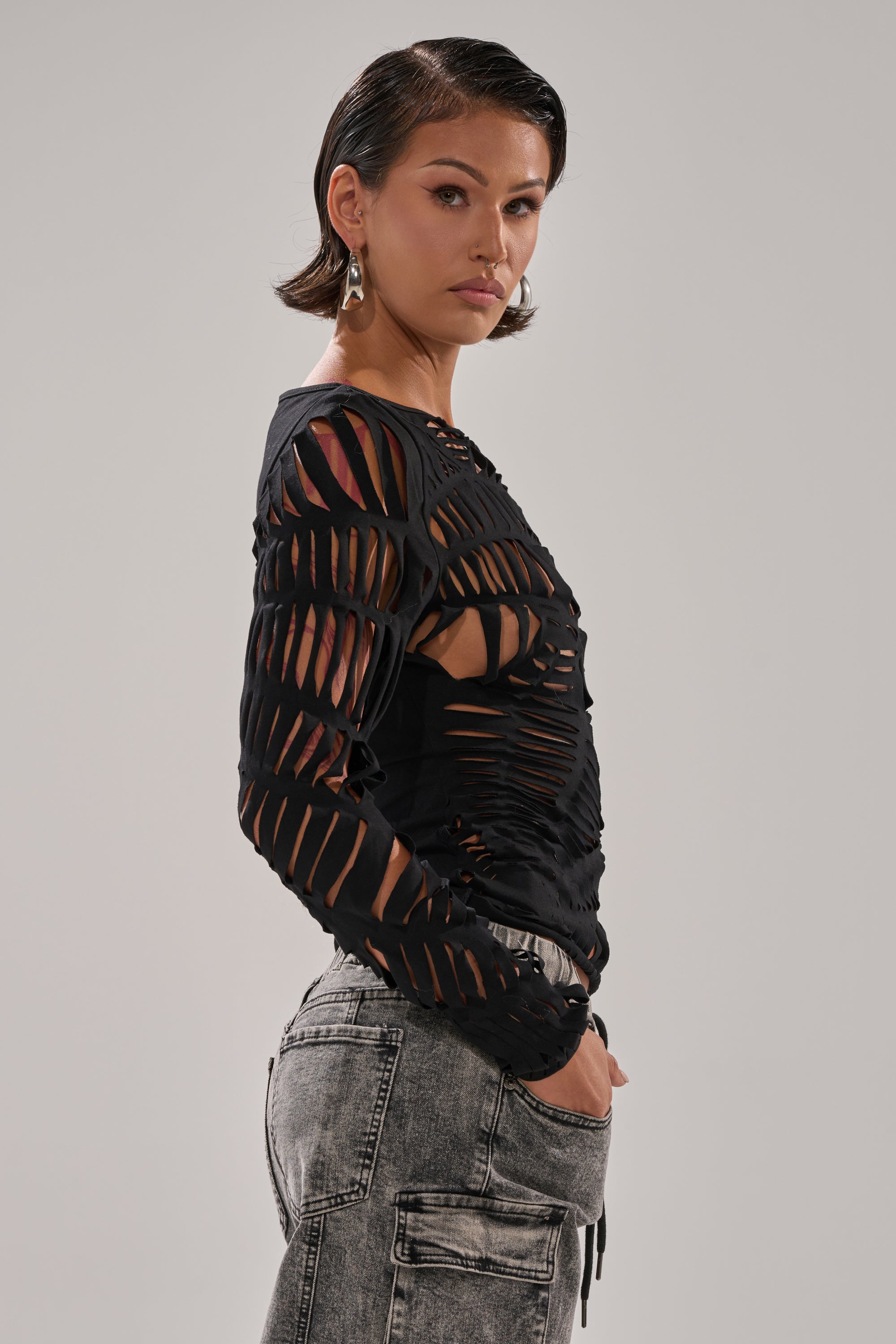 A woman with short brown hair wears the curve-hugging SPIDER WEB CUTOUT TOP in black and high-waisted acid-wash jeans. She stands sideways with hands in her pockets against a plain light background.