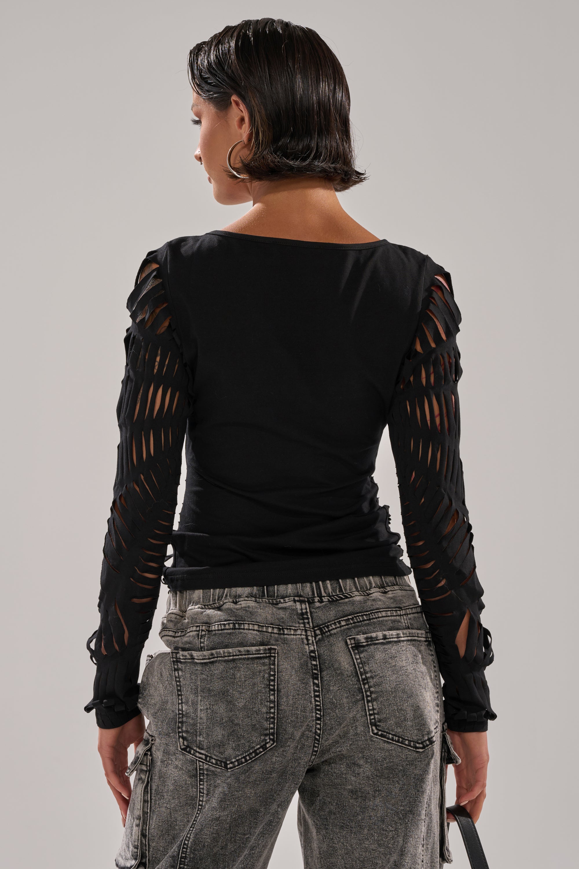 A woman with short dark hair, viewed from behind, wears the SPIDER WEB CUTOUT TOP featuring shredded cut details and acid-wash gray jeans with large back pockets, standing against a plain light gray background.
