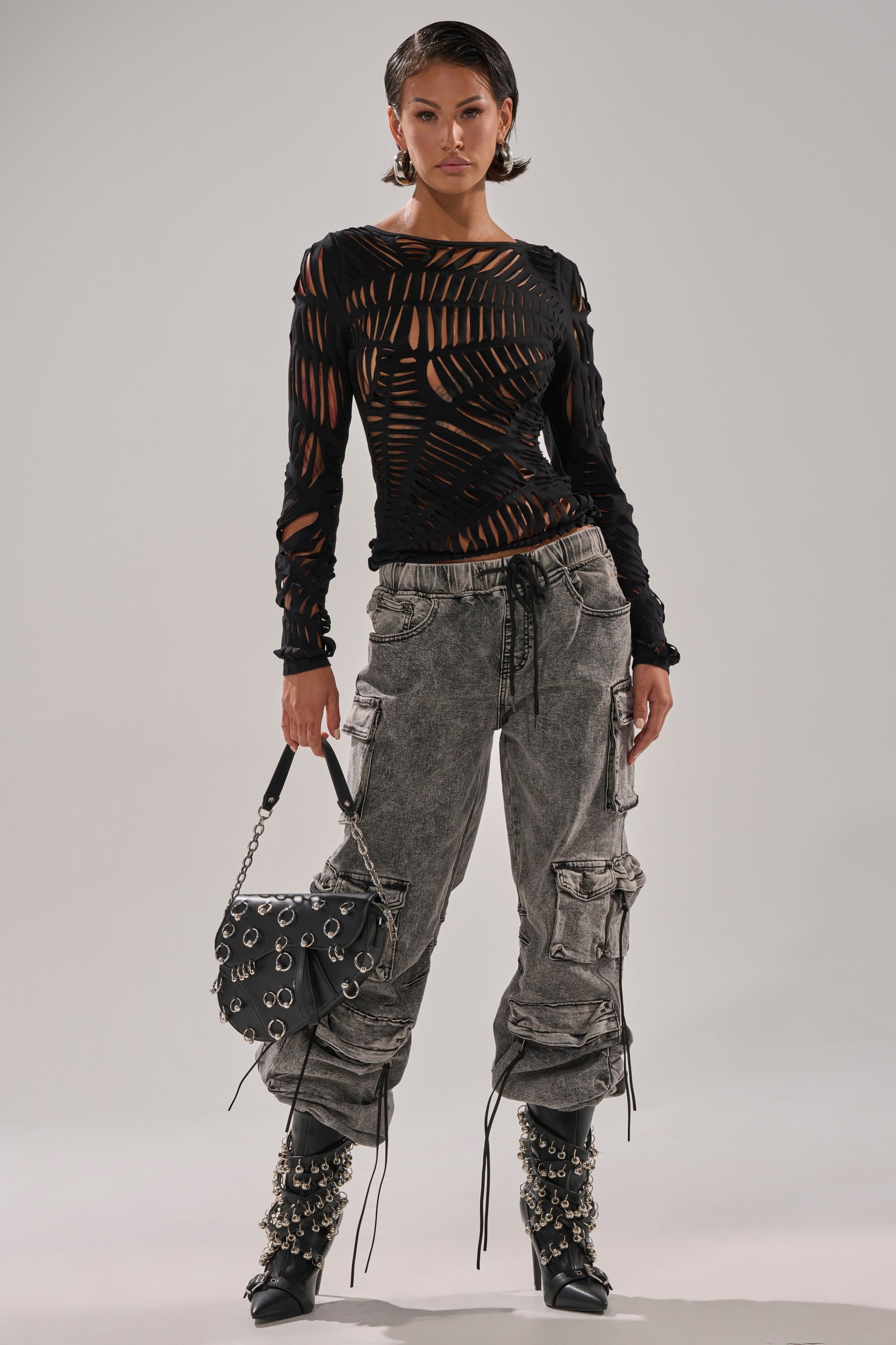 A person poses confidently in the SPIDER WEB CUTOUT TOP—black, long-sleeve with spiderweb cutouts—paired with light gray cargo pants, studded black boots, and a black handbag adorned with silver rings.