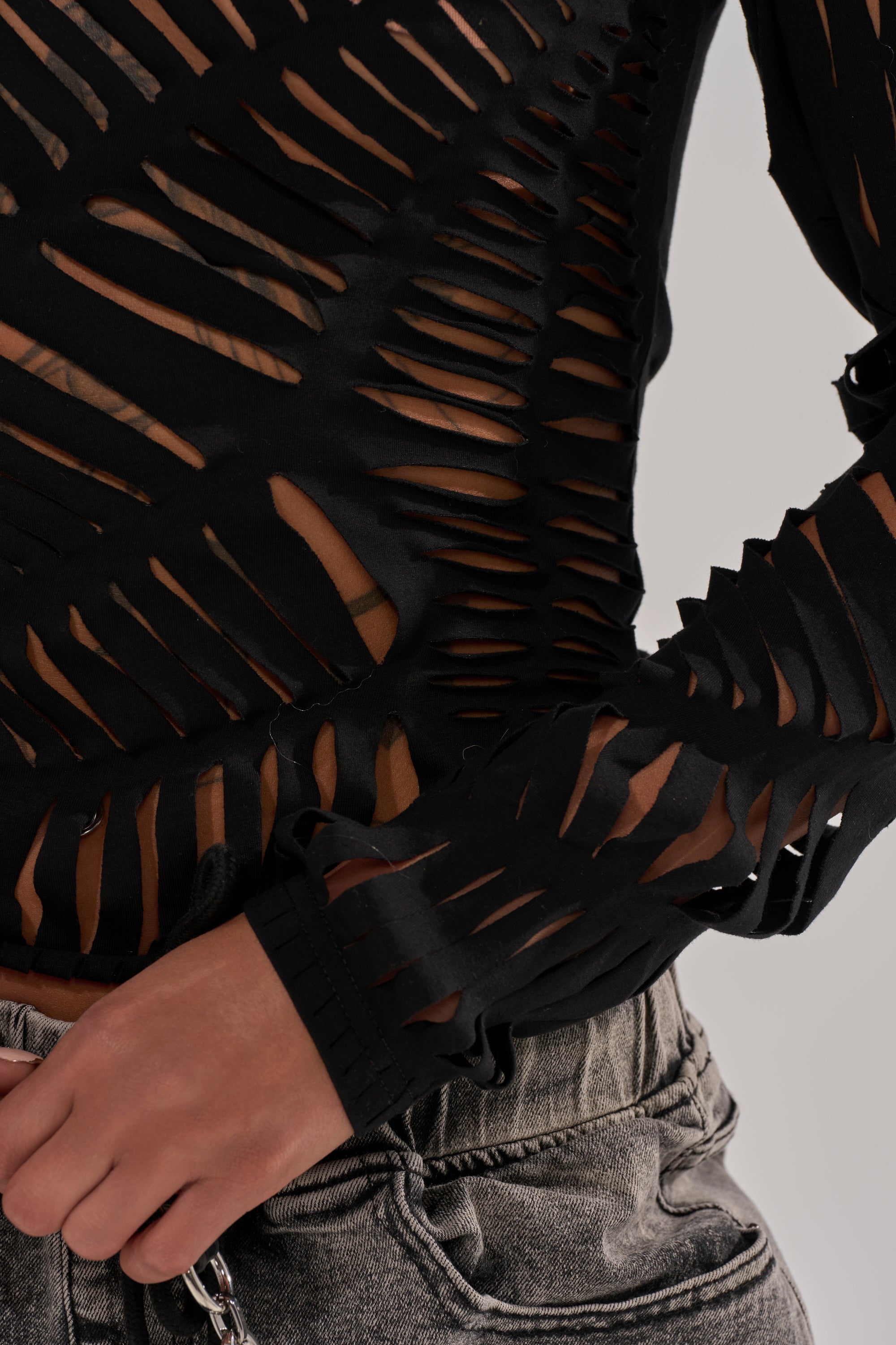 A close-up of a person in the black SPIDER WEB CUTOUT TOP, featuring shredded spider web details that reveal skin. They pair it with gray denim jeans and accessorize with a silver chain.