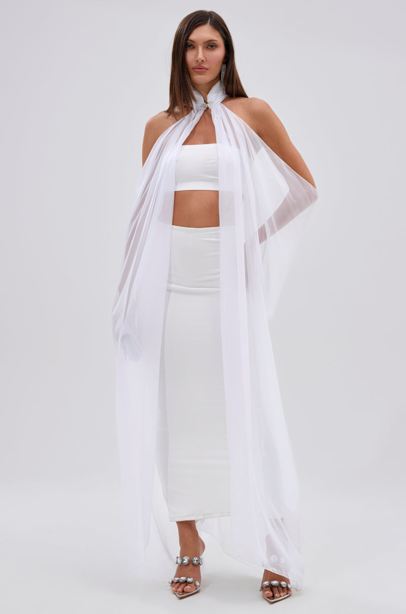 A woman wearing the GLOWING CHIFFON BLOUSE IN WHITE is styled with a strapless crop top, high-waisted skirt, and silver heels; her blouse cape drapes from neck to floor, and she has shoulder-length brown hair.