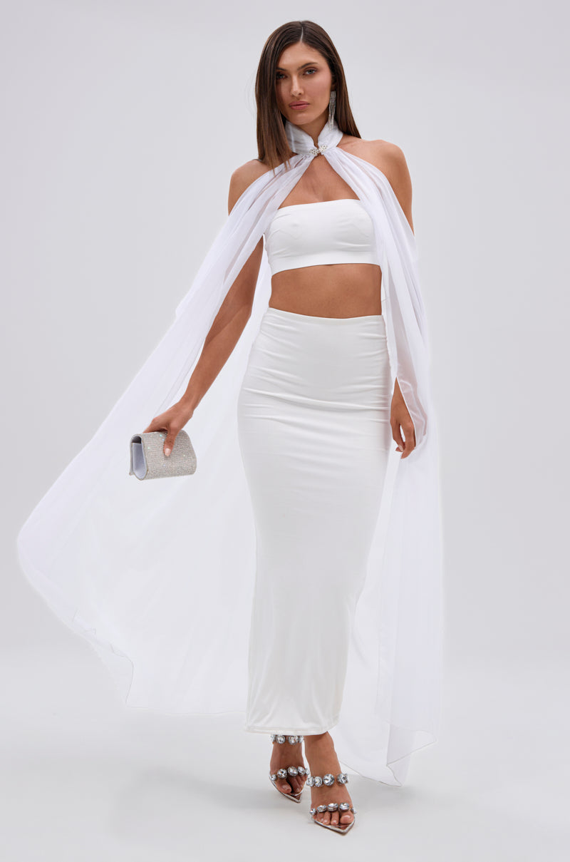 A woman wears the Glowing Chiffon Blouse in White, a sleeveless, fitted crop top paired with a matching skirt and sheer white cape. She accessorizes with a silver clutch and embellished sandals, standing before a plain light background.