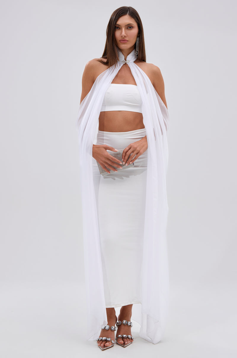 A woman stands barefoot, facing forward with hands clasped, wearing the Glowing Chiffon Blouse in White against a plain background, styled with a long white skirt and sheer cape.