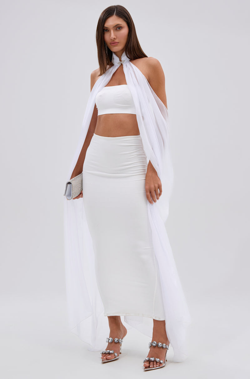 A woman styles the GLOWING CHIFFON BLOUSE IN WHITE as a cape over a white bandeau crop top and long skirt, finishing the look with embellished silver high-heeled sandals and a sparkling silver clutch.