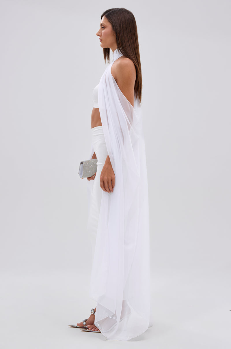 A woman in profile wears the Glowing Chiffon Blouse in White with a long, sheer cape, paired with a white skirt. She holds a silver clutch, wears silver heels, and her straight brown hair is down against a plain light gray background.