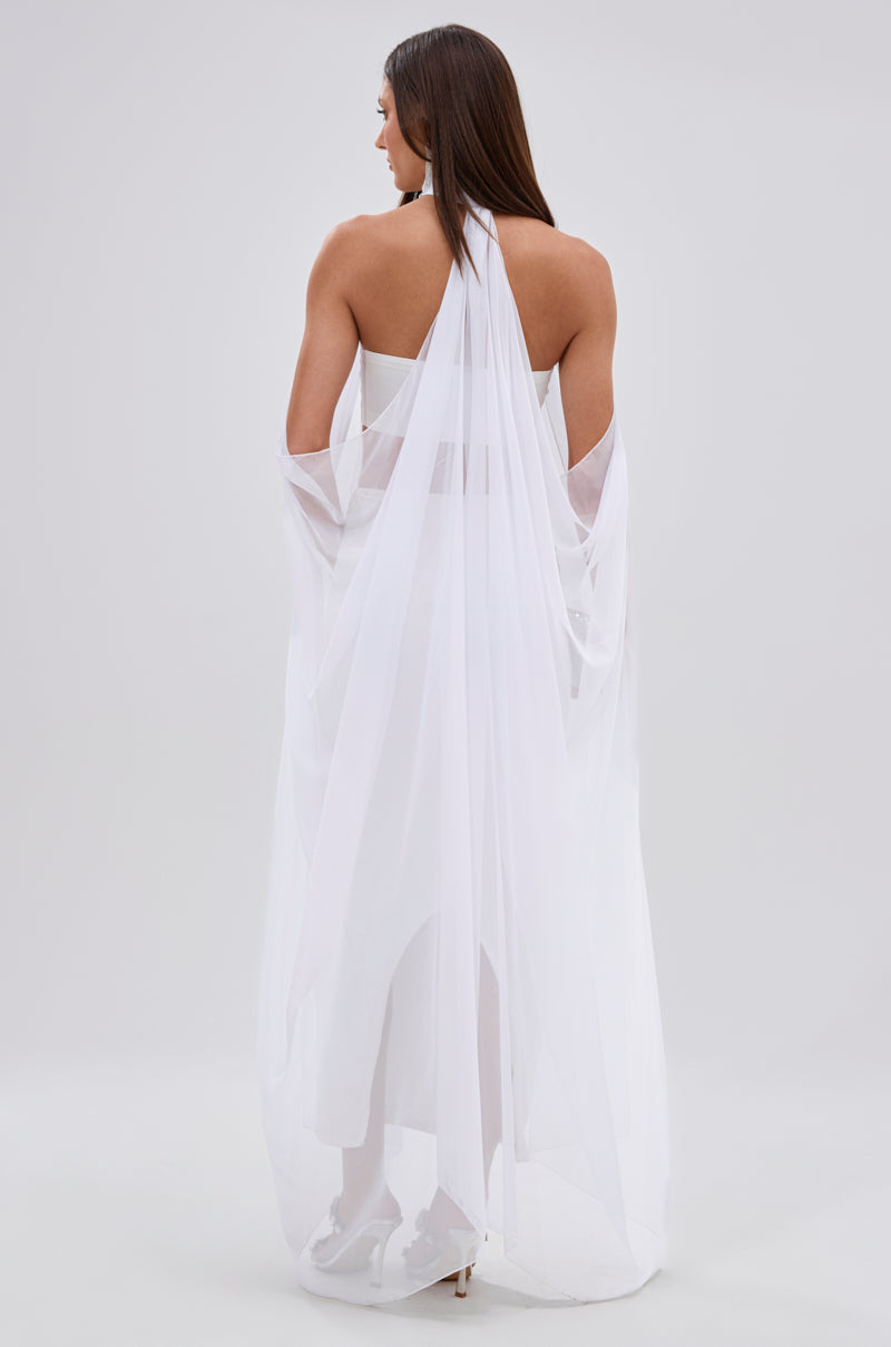 A woman stands facing away from the camera, wearing the GLOWING CHIFFON BLOUSE IN WHITE, which features a sleeveless design with flowing, sheer fabric. Her long hair cascades down her back against a plain light background.